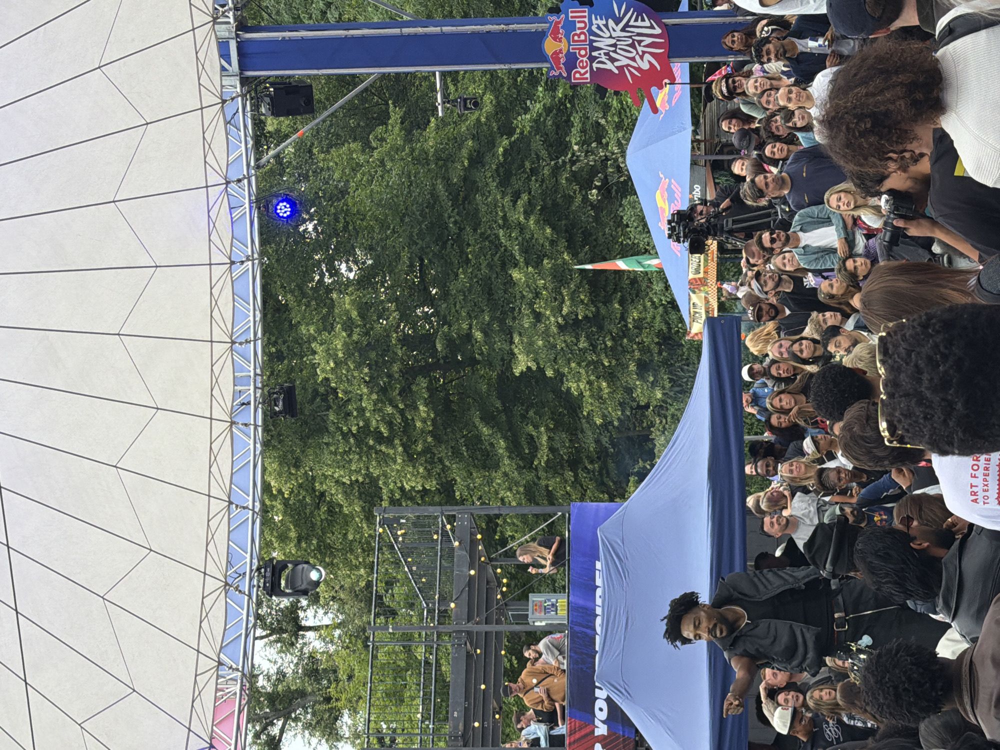 The image depicts an outdoor event under a large white canopy. In the foreground, a Black man with dreadlocks, wearing a black vest over a black t-shirt and black pants, is gesturing with his right hand towards the crowd. He appears to be a performer or host, speaking to the audience. Behind him, a large crowd of diverse people are gathered, many of whom are looking towards the performer.
In the background, there are several tents, including a blue tent with a Red Bull logo and a "Dance Your Style" graphic. There is also a raised platform with people standing on it, some with drinks, and string lights are visible. Trees and greenery form the backdrop to the scene. A Red Bull flag is also visible. A camera on a tripod is positioned behind the crowd, facing the stage area. The overall atmosphere suggests a lively, public event. The Red Bull branding and the "Dance Your Style" graphic suggest this is likely part of a Red Bull sponsored dance competition or festival, which are often held in public spaces and can take place in cities like Amsterdam, Netherlands.