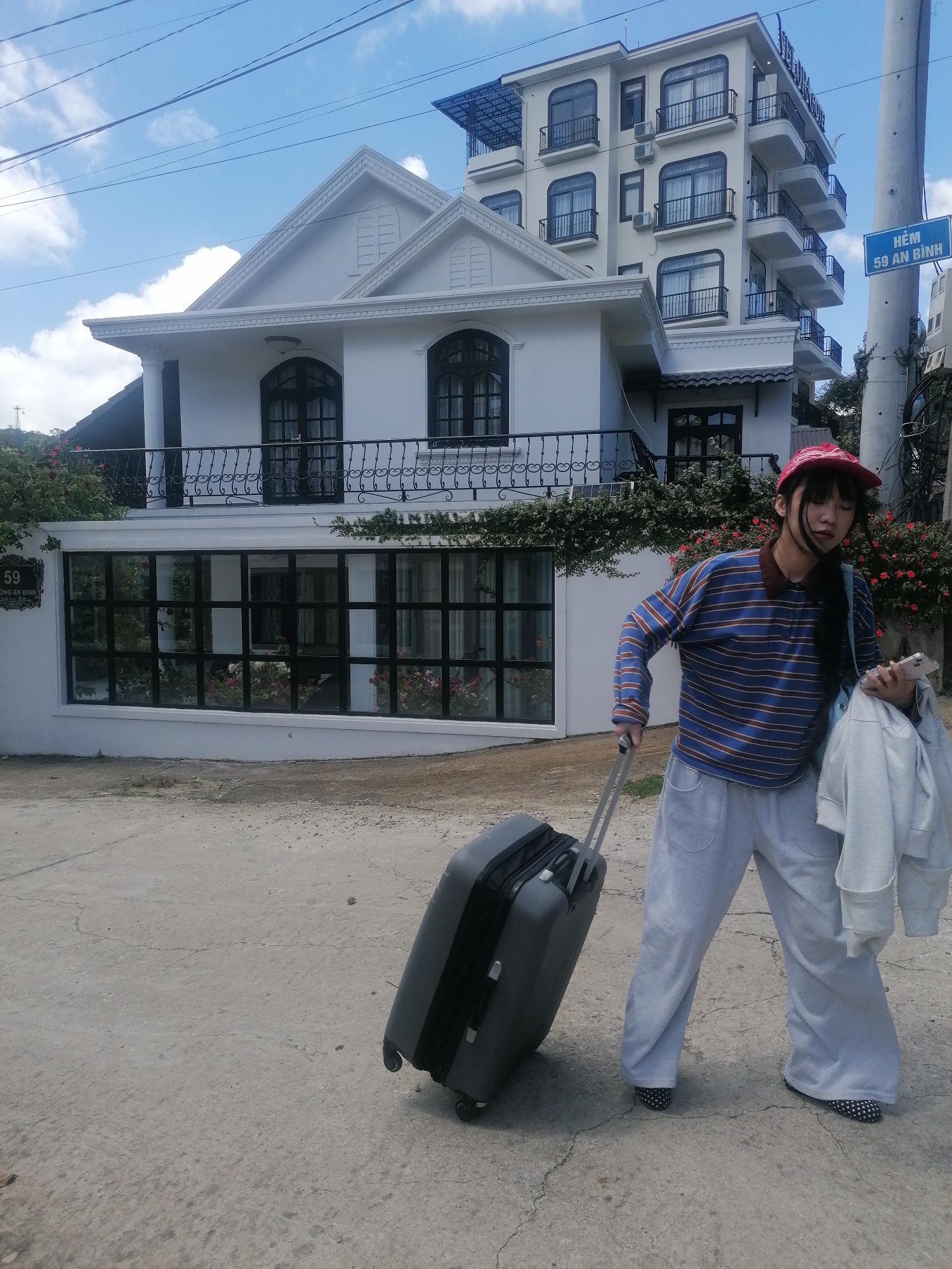 A young Vietnamese woman, wearing a red baseball cap, a blue and brown striped long-sleeved shirt, and light gray sweatpants, stands on a cracked asphalt road in Da Lat, Vietnam. She is pulling a dark gray suitcase with a silver handle and holding a white jacket over her right arm, with a white smartphone in her left hand. Behind her is a two-story white house with a balcony and large windows. To the right of the house is a taller, multi-story building with balconies and arched windows. A blue street sign with white text that reads "HẺM 59 AN BÌNH" is visible on a utility pole on the right side of the image. The sky is blue with scattered white clouds.