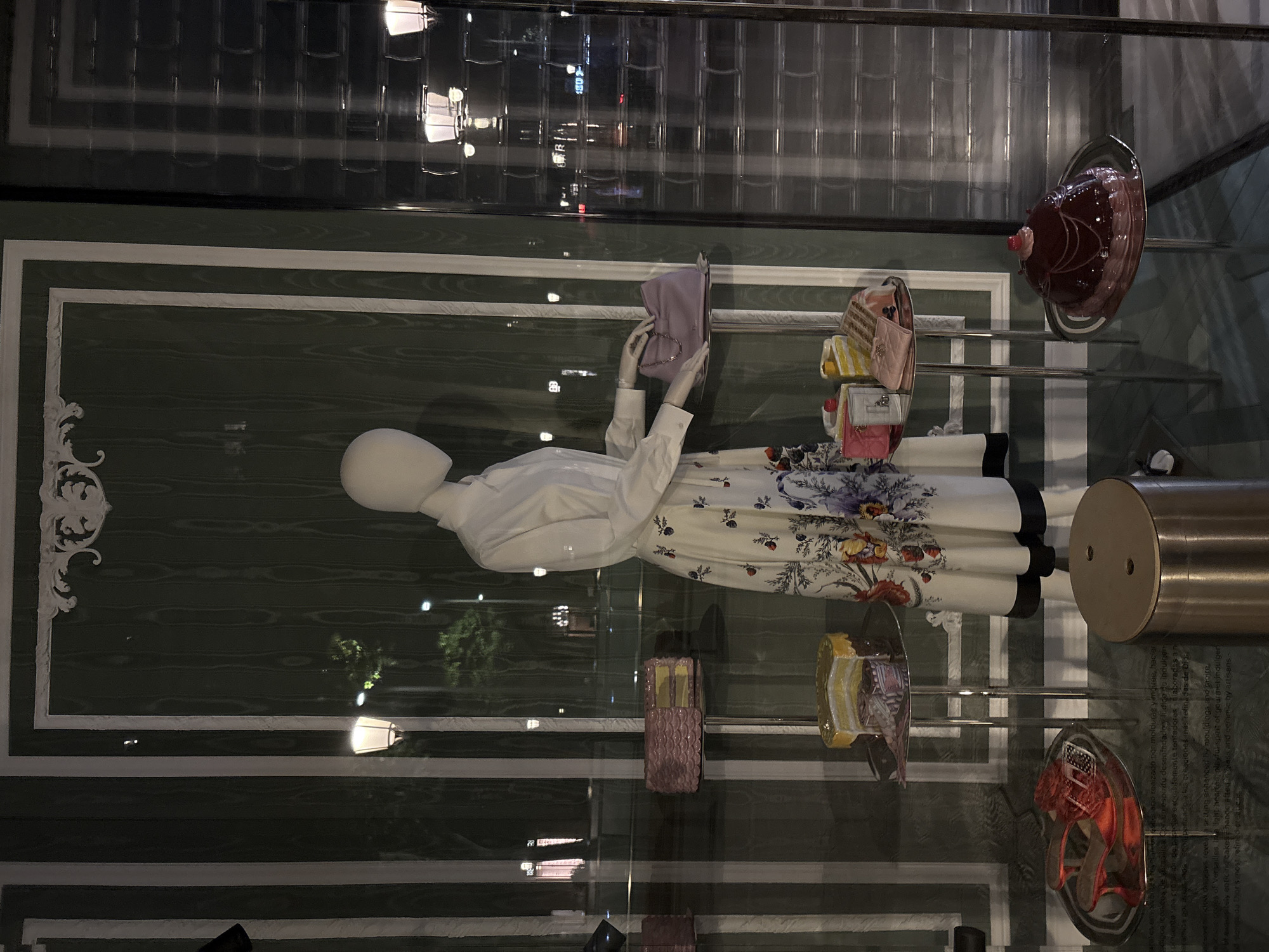 The image depicts a window display, likely in a high-end retail store in Barcelona, Spain, given the presence of Spanish text. The main subject is a mannequin dressed in a white blouse and a long, floral-print skirt. The mannequin appears to be holding a light purple bag. The display includes other items such as cakes and handbags that are placed on small shelves, creating a layered effect. The background consists of a decorative green panel with white trim, reminiscent of Parisian design. The window reflects a city scene outside, with street lights and building facades. The display suggests a theme of luxury, fashion, and perhaps a celebration of baked goods.
