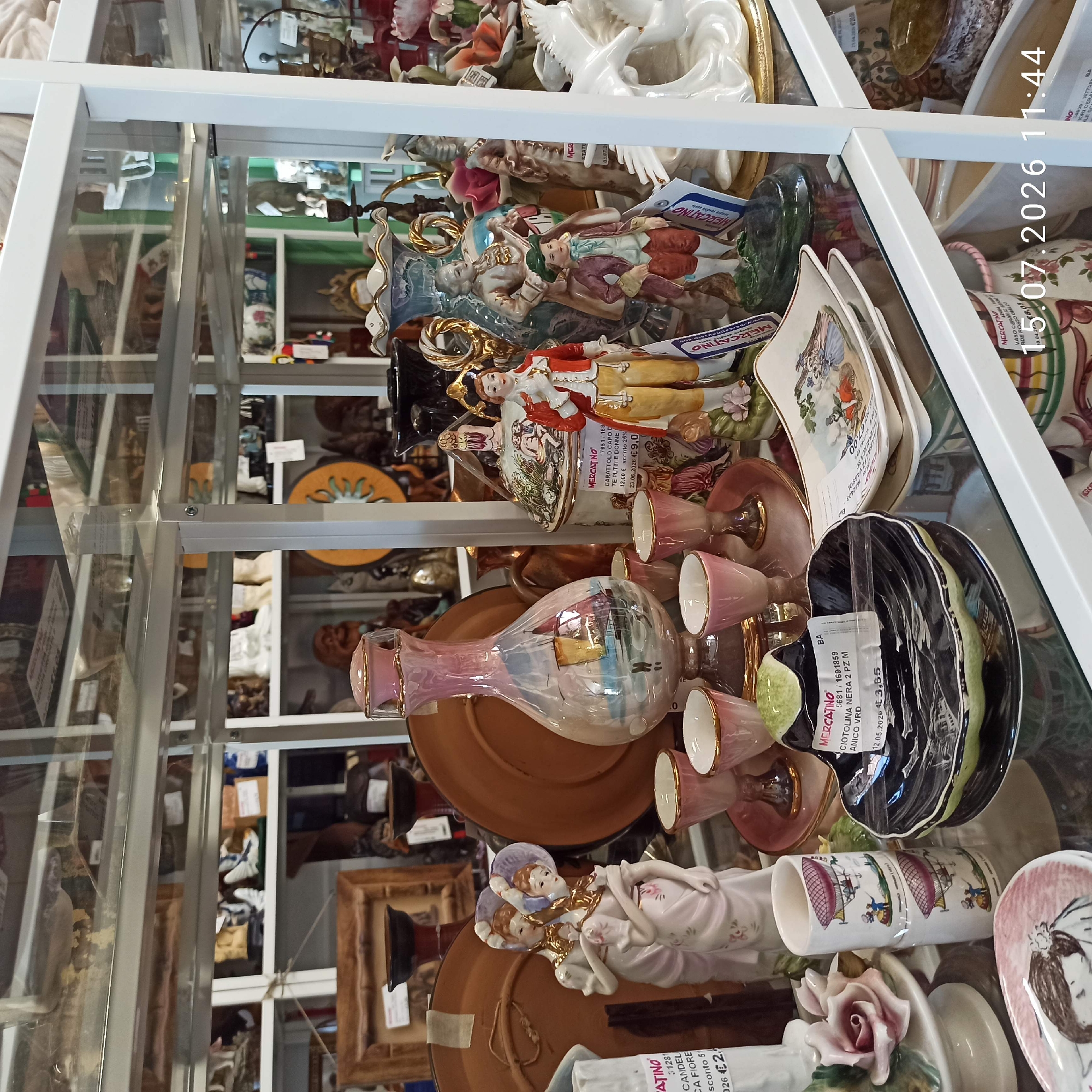 The image shows a retail display of antique or decorative ceramic items on glass shelves. The items include figurines of people in historical clothing, vases, plates, and cups, many with ornate details. There is a particular focus on the lower shelves, where several pieces are prominently displayed. On the left side, a porcelain figurine of two women in pink dresses with hats stands near a stack of two cylindrical cups decorated with hot air balloons. To the right of these cups, a black and green ceramic dish with a wavy edge is visible, bearing a price tag that reads "MERCATINO CIOTOLINA NERA 2 PZ M ANICO VRD 12.05.2026 €3,65". Further to the right, a small decorative plate with a scene of people on a bicycle is stacked on another plate. In the background, more ceramic items and glassware are visible on higher shelves. The overall impression is that of an antique shop or a curated display of collectibles. The timestamp overlay indicates "15.07.2026 11:44," suggesting the image was taken on this date and time, though it is a future date. Without additional context, it is impossible to definitively link this image to Molfetta, Italy, although the presence of Italian price tags suggests it may be a location in Italy.