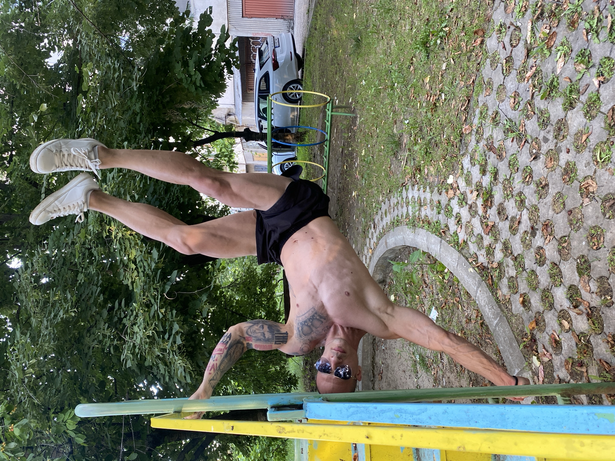 An athletic adult male is depicted performing a horizontal body flag exercise on a playground structure in what appears to be an urban park or courtyard in Pleven, Bulgaria. The man, shirtless, exhibits a muscular physique with visible veins and extensive black and grey tattoos covering both arms and extending onto his torso. He has a shaven head and wears reflective sunglasses, black athletic shorts, and white sneakers.
He is positioned horizontally, parallel to the ground, with his body supported primarily by his left hand gripping a vertical light-green bar of a multi-colored playground apparatus, which also features yellow and blue bars. His legs are extended upwards, maintaining the horizontal posture, while his right arm is extended downwards towards the ground, contributing to balance. His head is tilted upwards, looking towards the viewer. He wears a black wristband on his right wrist.
The ground beneath him consists of a section of grass interspersed with scattered dry leaves and a path paved with hexagonal concrete blocks, each containing a circular opening for vegetation. A curved concrete curb separates the paved area from the grass. In the background, numerous deciduous trees with dense green foliage are visible. Beyond the trees and in the mid-ground, a white Skoda Octavia station wagon is parked, along with a partial view of another light-colored vehicle. Apartment buildings with visible facades and a reddish-brown door are present further in the background. A low green playground structure with three hoops (yellow, blue, yellow) is visible on the grass in the mid-ground, behind the man's legs. The scene is illuminated by natural daylight.
