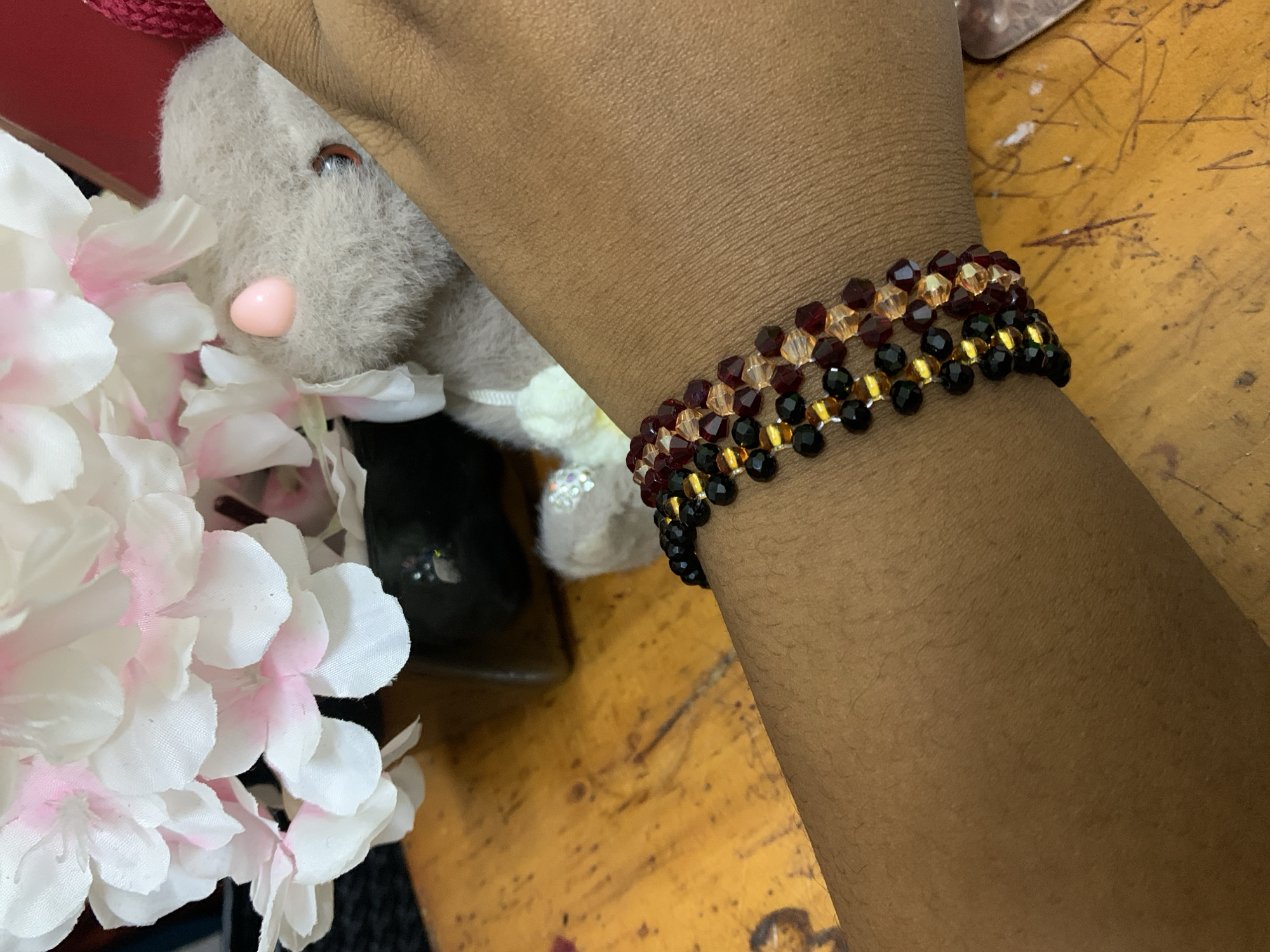A close-up shot focuses on a person's wrist, adorned with a beaded bracelet featuring rows of dark red, amber, and black beads. The person's skin tone is brown. Adjacent to the wrist, partially visible, are white artificial flowers with pink accents and a stuffed toy rabbit. The background consists of a wood-like surface with visible markings. The image was taken in a room and is not obviously or necessarily linked to Dar es-Salaam, Tanzania.