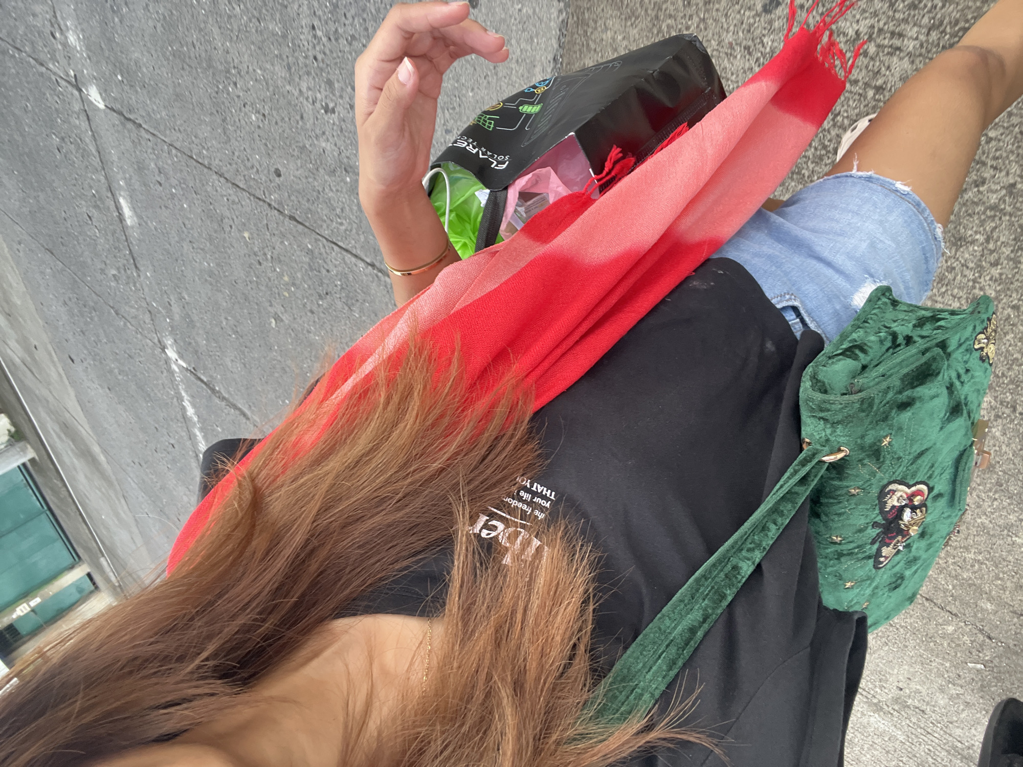 A first-person perspective shot shows a person standing on a gray paved surface. The person is wearing denim shorts and a black t-shirt, carrying a red scarf, a green velvet purse with embellishments, and a black shopping bag. The bag has "FLARE" written on it. The person's right hand is visible. The scene takes place on a city street, with a building visible in the background. The specific location within Davao City is not discernible from the image.