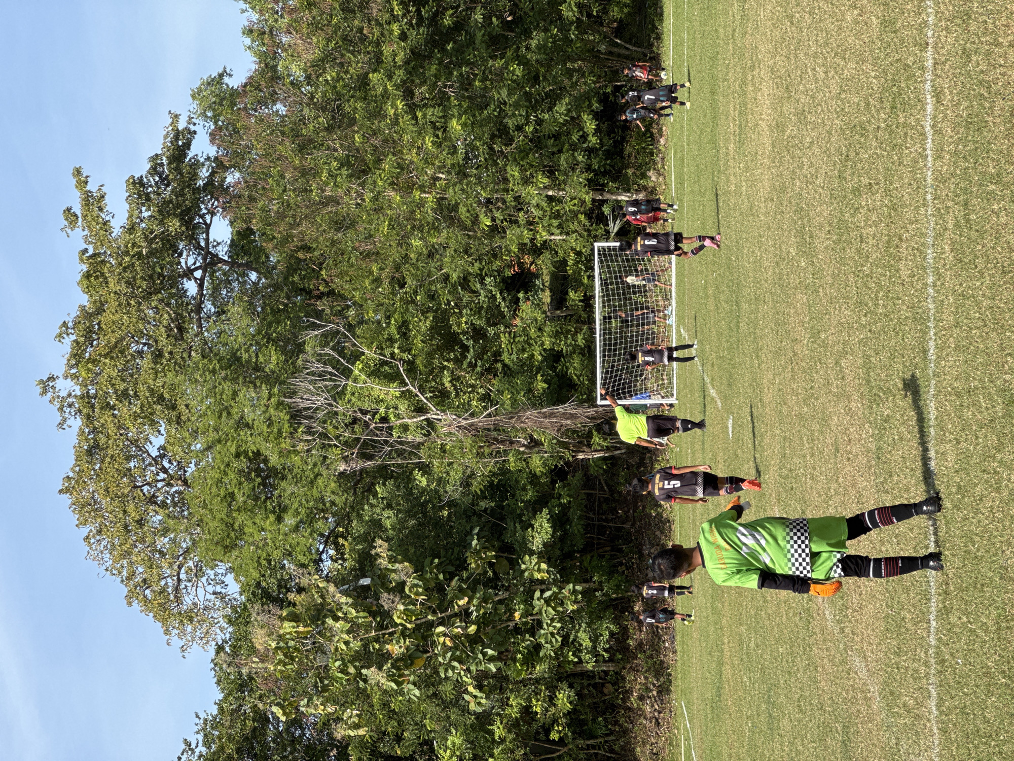 The image depicts a youth soccer game in progress at a field located in Kuta Selatan, Indonesia. The playing surface is grass, with white lines marking the boundaries. A soccer goal with a net is visible, with players positioned around it. One player is wearing a green and black checkered jersey, black shorts, black and red socks, and black shoes. Other players are wearing black jerseys. The environment includes trees lining the edge of the field, and a clear blue sky is visible in the background.