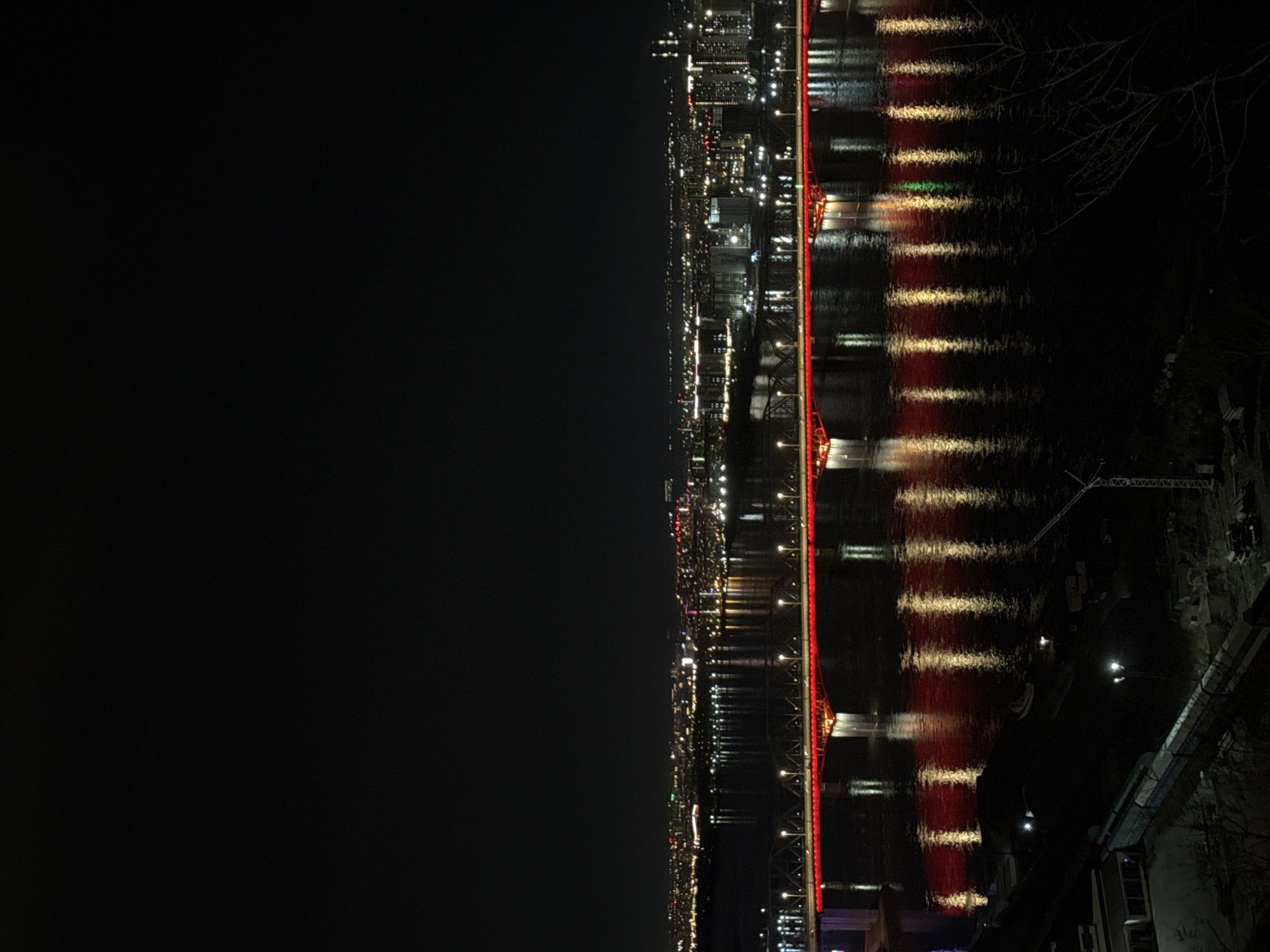 An eye-level, rotated, horizontally-oriented, nighttime photograph shows a view of the city of Krasnoyarsk, Russia. The city lights stretch along the right side of the image, with a body of water reflecting the lights from the city. A bridge, lit with red lights, crosses the water. To the right, some foliage is present with some buildings at the lower right. A vast black area fills the left side of the image, representing the night sky or unlit land.