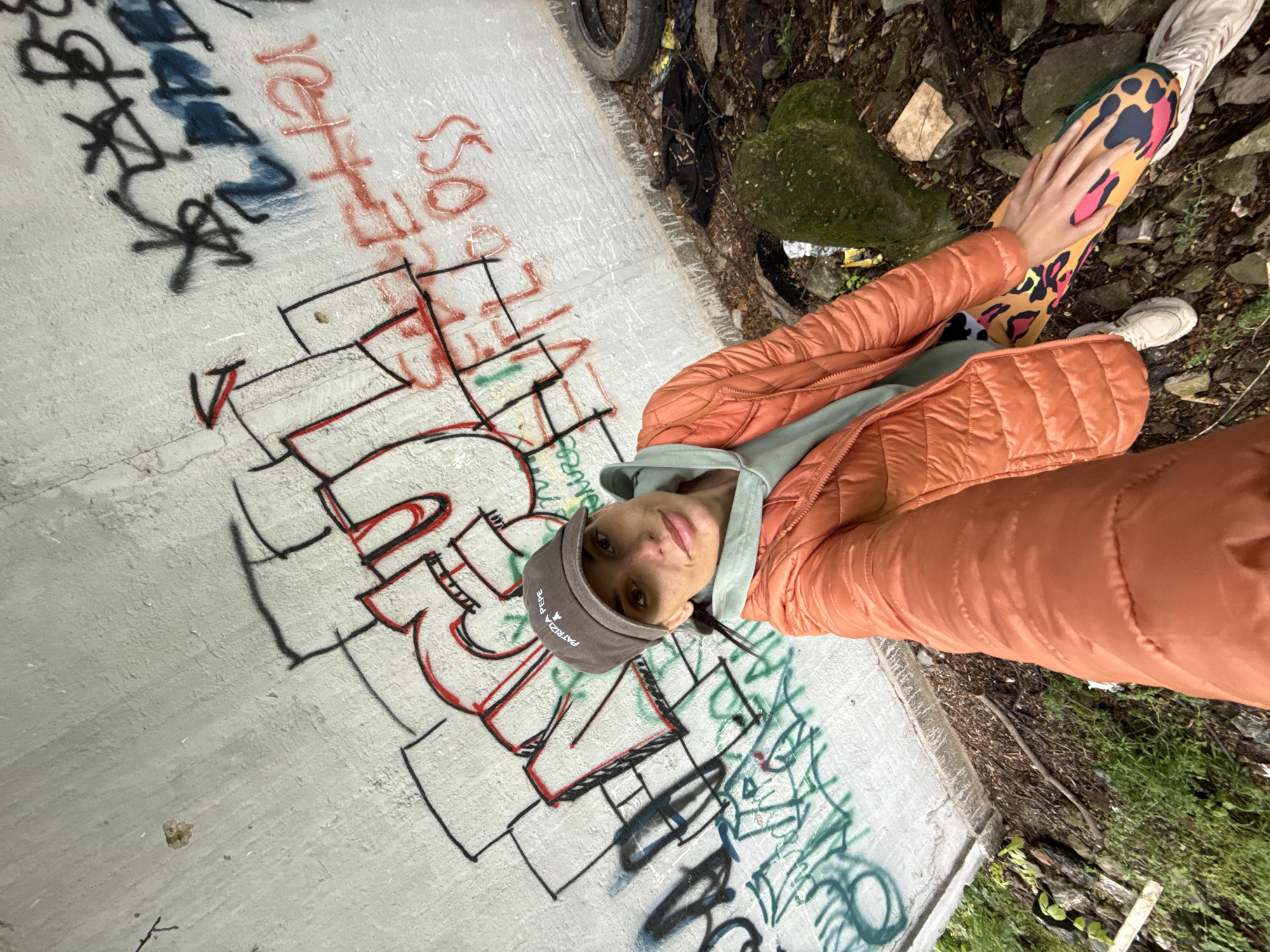 A woman wearing an orange puffer jacket, a light green hoodie, a grey "Patrizia Pepe" baseball cap, and leopard-print leggings is posing for a selfie. She is standing on a concrete structure covered in graffiti. The graffiti includes lettering in black, red, blue, and green. A tire, trash, moss, and rocks are visible near the edge of the structure. Vegetation is present.
I don't have information about the precise coordinates in the city of Pleven, Bulgaria, for the presented image.