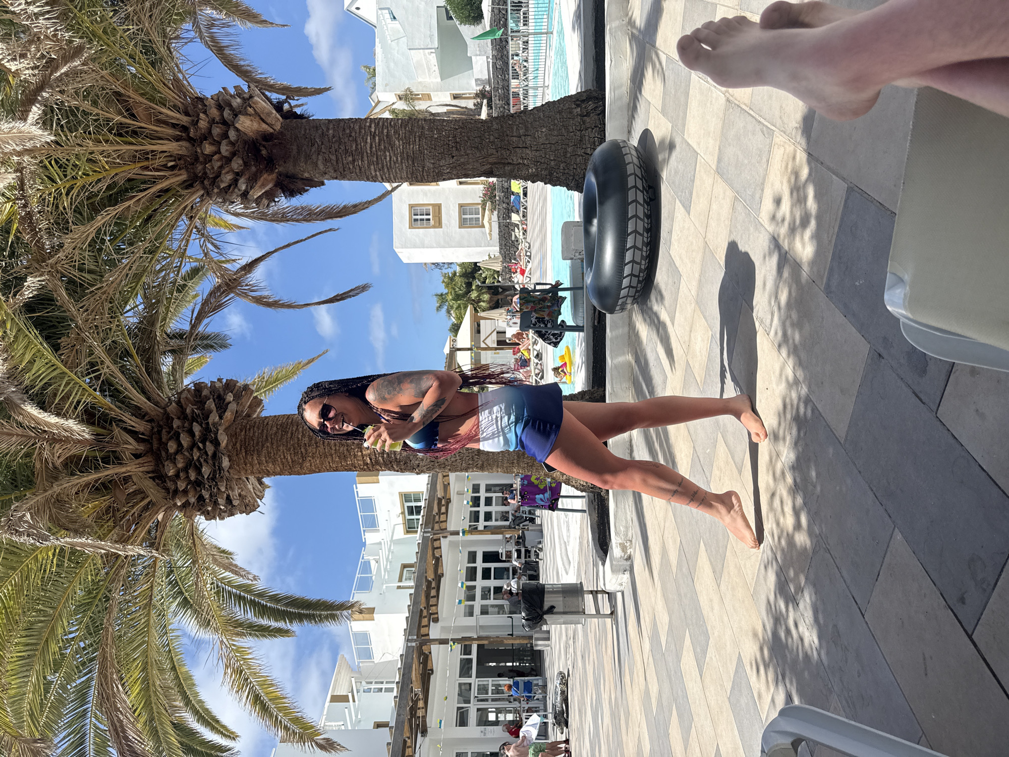 The image depicts a sunny outdoor scene in Puerto del Carmen, Spain. A woman in a swimsuit stands on a tiled surface beside a palm tree, holding a beverage. Behind her is a multi-story white building with windows and balconies, and a glimpse of a swimming pool with people and lounge chairs can be seen. An inflatable tube rests against the palm tree trunk. In the upper right corner of the frame, a pair of human feet suggest the presence of someone relaxing nearby.