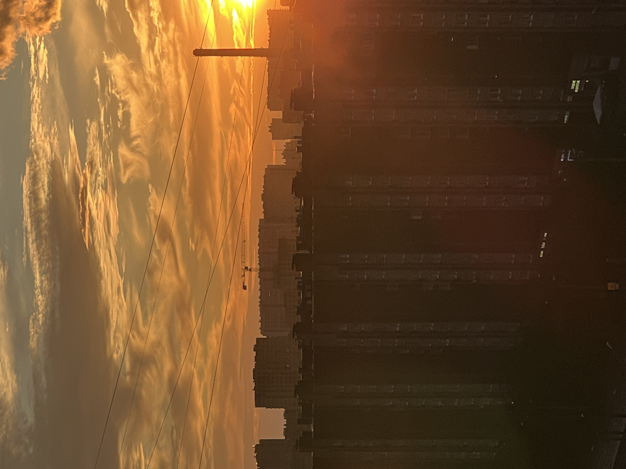 A vertical, full shot captures a city skyline at sunset. The sky is ablaze with vibrant orange and yellow hues, streaked with wispy white clouds. Several power lines crisscross the frame horizontally. In the distance, silhouettes of apartment buildings and a tall smokestack are visible against the glowing sky. The foreground is dominated by the dark, towering facades of more apartment buildings, their windows appearing as small, rectangular shapes. The overall atmosphere is one of urban twilight, with the warm light of the setting sun casting long shadows and silhouetting the structures.
