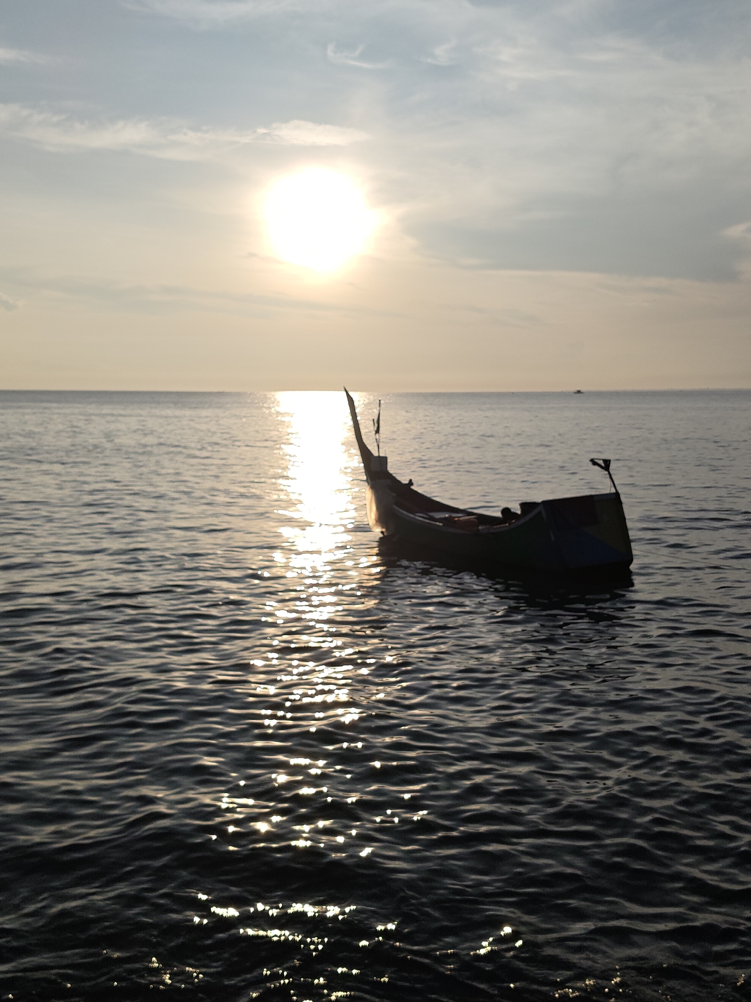 The image depicts a solitary boat silhouetted against the backdrop of a sunset over the ocean. The sun, a bright orb, is positioned high in the sky, its light reflecting off the water's surface, creating a shimmering path towards the viewer. The boat, appearing dark in contrast to the bright sky and water, is anchored or adrift in the calm sea. The water is marked by gentle waves, which add texture to the scene. The sky is partially covered with light clouds. The location is the city of Lhokseumawe, Indonesia.