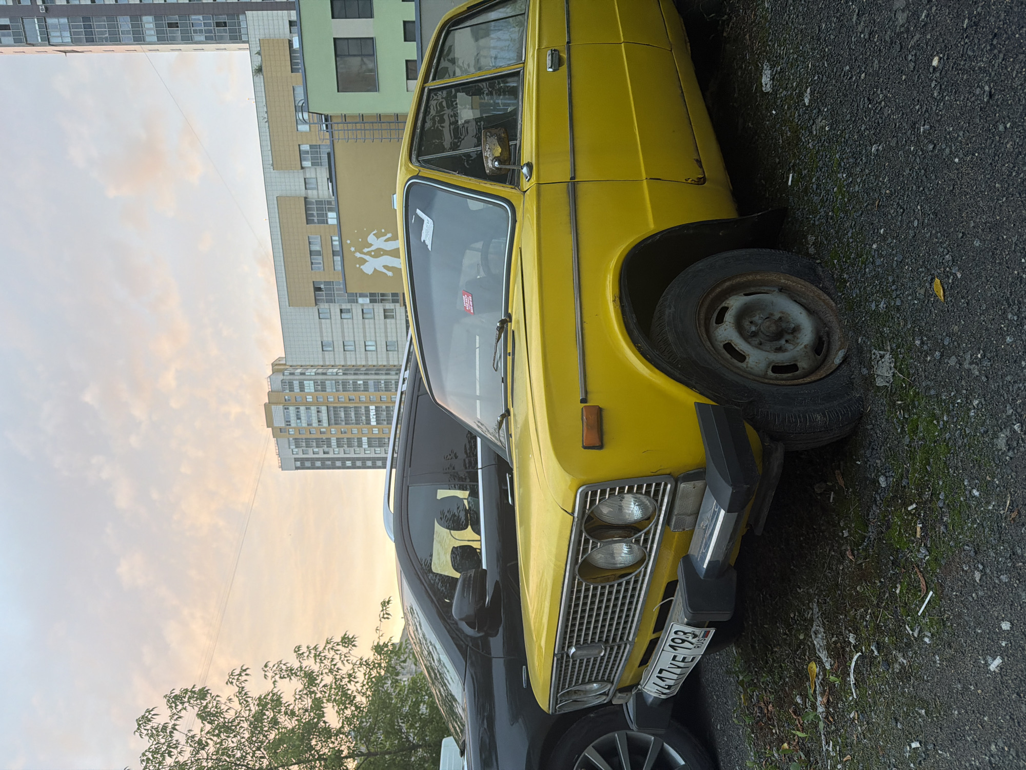 This is a medium shot of a bright yellow vintage sedan parked next to a dark-colored SUV in an urban setting. The yellow car is in the foreground, taking up most of the right side of the frame. It has a chrome grille, two round headlights, and a visible license plate that reads "K417VE 193 RUS". The car appears to be parked on a gravel surface, and the front passenger-side tire is flat. The SUV is visible to the left of the yellow car, with its side and rear window in view. In the background, several modern apartment buildings rise against a pale, cloudy sky with hints of sunset colors. The buildings are a mix of architectural styles and colors, including yellow, beige, and light green. A tree with green foliage is on the far left of the frame, partially obscuring the sky and buildings. The overall scene suggests a residential area in a city, possibly in the late afternoon or early evening.