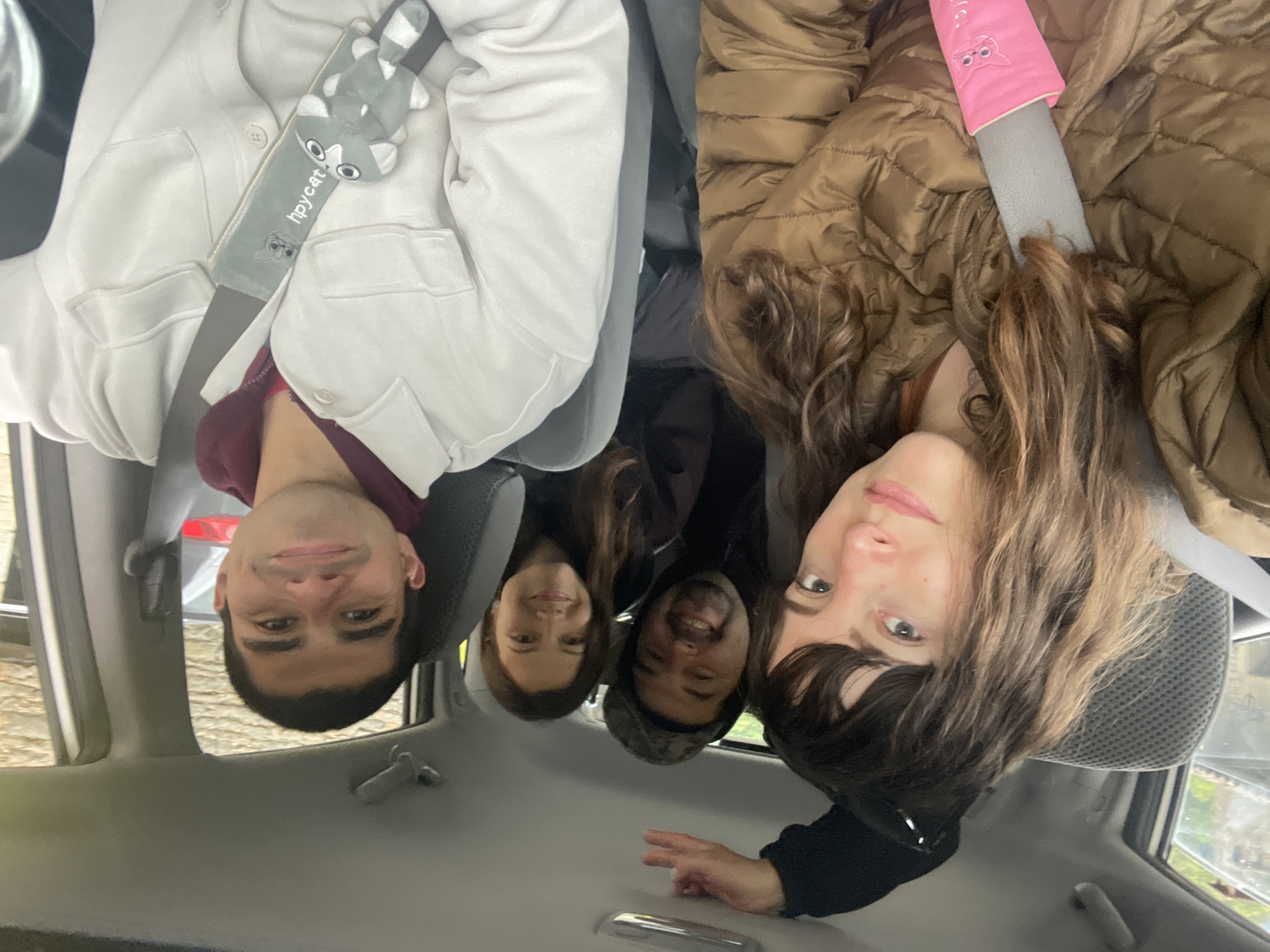 The photograph depicts four individuals inside a car, all facing the camera. The image is oriented upside down. The person on the left wears a light-colored jacket over a maroon-colored top and has a gray cat-shaped seatbelt cover. Next to them is a woman with long brown hair, followed by a man with short dark hair. The person on the right wears a brown puffer jacket with a pink seatbelt cover. All four individuals are wearing seatbelts. The car interior is visible, showing the dashboard, seats, and door panels. Through the car windows, a glimpse of the outdoors can be seen, likely within the city of Rupit, Spain.