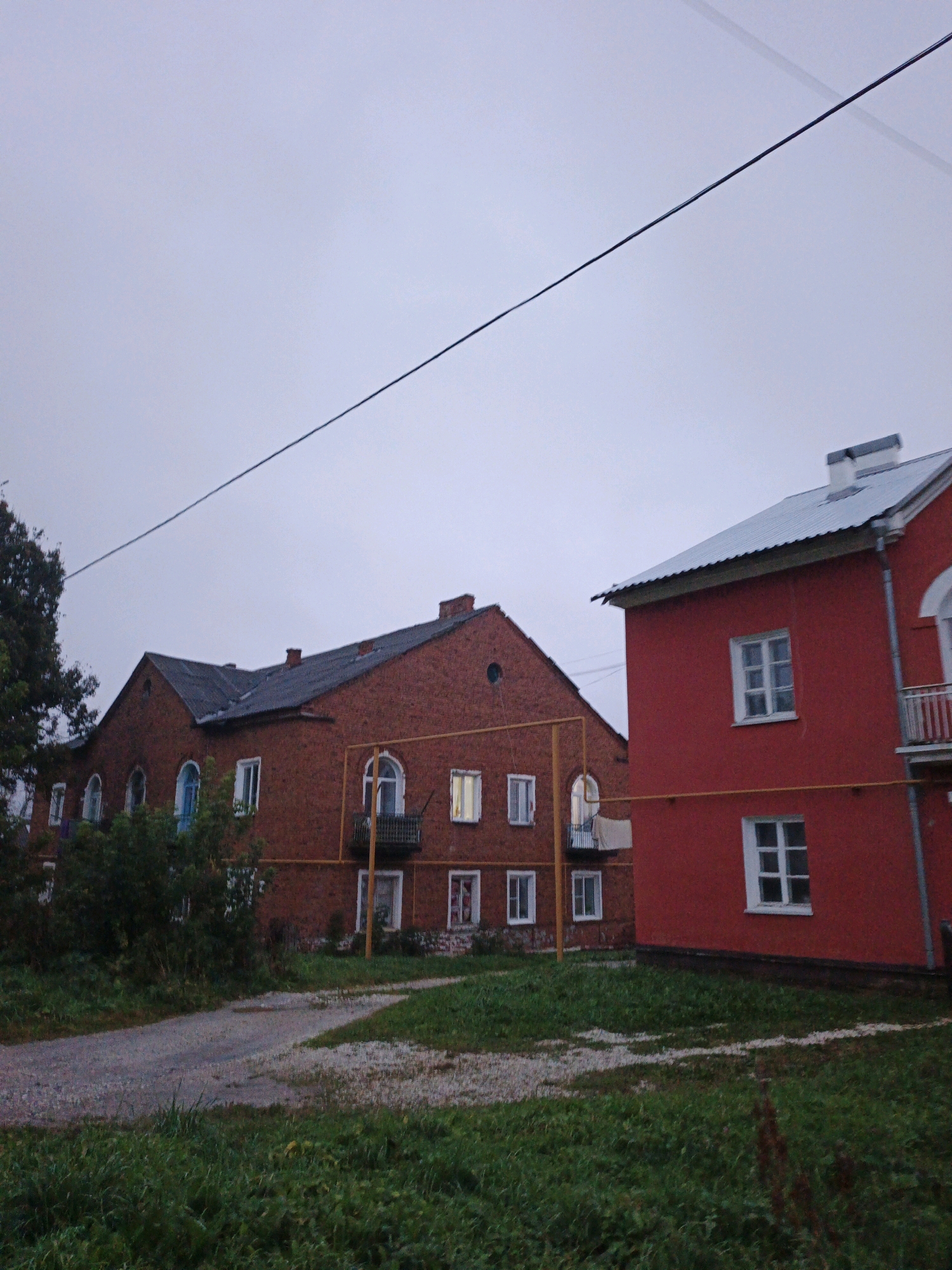 The image shows two apartment buildings in Medvedyovka, Russia. The building on the left is made of red brick and has a dark gray roof. It features several arched windows and balconies. The building on the right is painted bright red and has a silver-colored metal roof with a white chimney. It also has several windows, some of which are multi-paned. A network of yellow pipes runs horizontally across the facade of both buildings. A single black wire stretches diagonally across the overcast sky. In the foreground, a gravel path curves through overgrown green grass. The scene appears to be a residential area on an overcast day.
