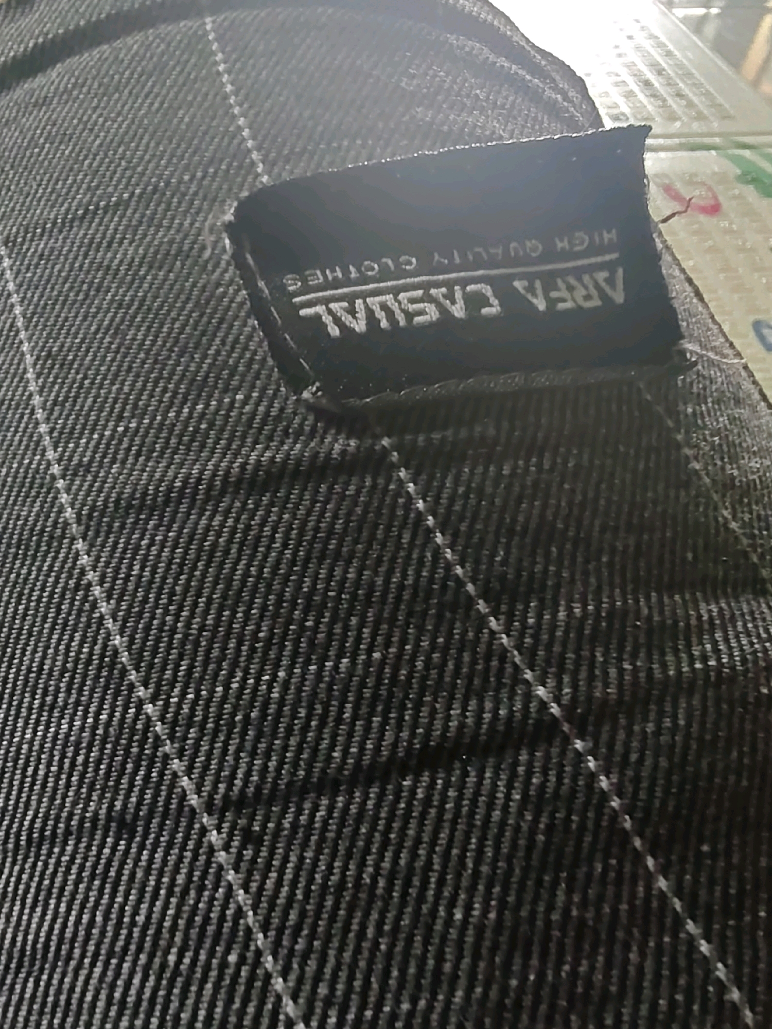 The image is a close-up of a dark fabric with a black label attached. The label has white text that reads "ARFA CASUAL HIGH QUALITY CLOTHES". The fabric appears to be denim or a similar material, with visible stitching lines. The lighting suggests an indoor environment, possibly a retail setting or a personal collection. There are no people, animals, or any clear indicators of a specific location in Sumberwono, Indonesia, present in the image.