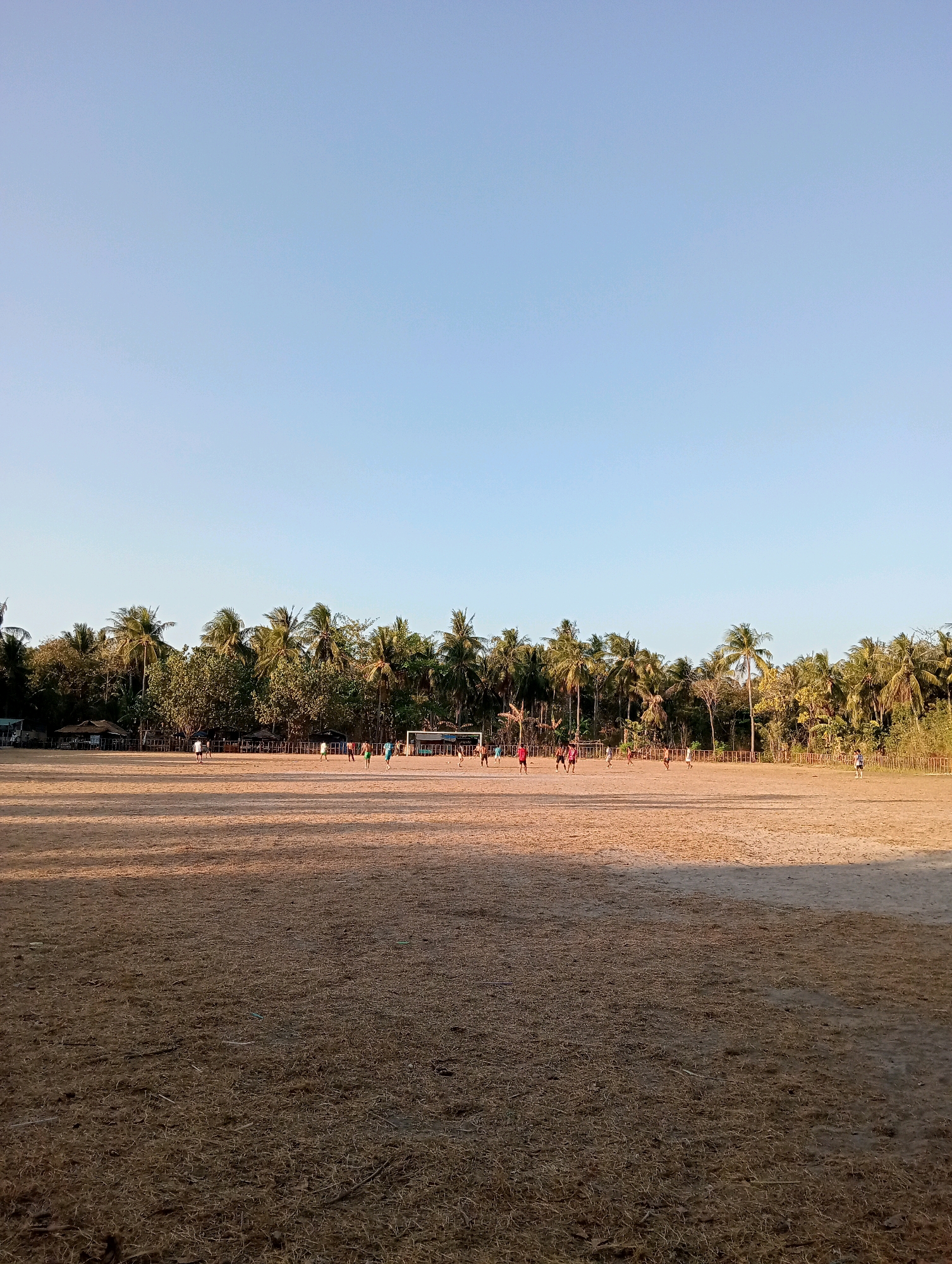 An outdoor, wide shot captures a sandy field with sparse, dry grass and a soccer goal at the far end. A group of individuals, appearing to be playing soccer, are visible on the field. A dense line of palm trees and other foliage forms the backdrop, with a clear blue sky overhead. In the foreground, the ground is covered in dry, straw-like grass, casting long shadows from the low-lying sun. A few structures, possibly small buildings or stalls, are situated among the trees on the left side of the image. The overall scene suggests a casual sporting event in a tropical environment.