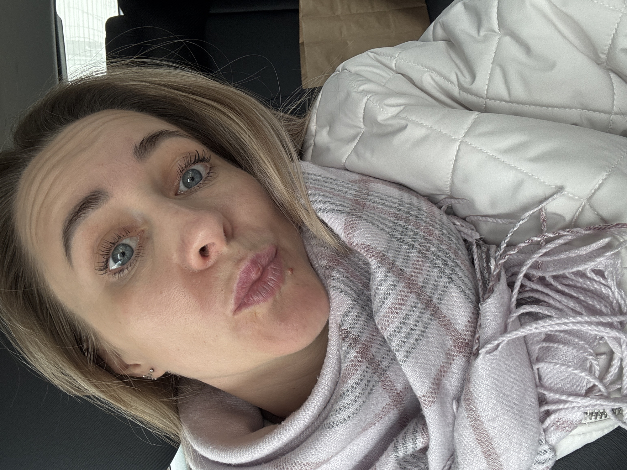 A woman with blonde hair and blue eyes is looking at the camera with pursed lips. She is wearing a light pink and gray plaid scarf and a white quilted jacket. The background is dark and out of focus, but appears to be the interior of a car or other vehicle. There are no clear landmarks or other information to suggest the location of the image is Chaikovsky, Russia.