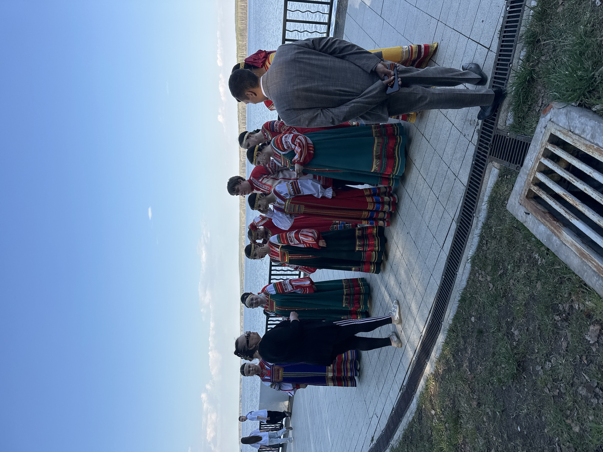 A group of people in traditional Russian folk costumes stand in a line on a paved surface near a body of water. A man in a suit stands facing the group, possibly taking a photograph. Another person in athletic wear stands near the end of the line of costumed individuals. The location is likely in the city of Chaikovsky, Russia, given the cultural context of the clothing. A metal grate borders the pavement. The sky is clear and blue.