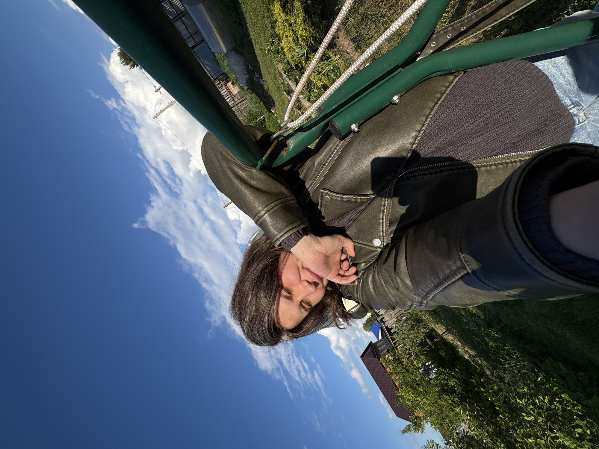 A woman is captured in an outdoor selfie, looking towards the camera with a slight smile and partially closed eyes, possibly due to bright sunlight. She has dark hair and is wearing a dark, possibly olive-green, leather-like jacket over a dark, ribbed knit top and blue jeans. Her right hand is raised, partially covering her chin and mouth.
In the foreground, a prominent green metal structure, which could be part of a swing set or garden frame, diagonally crosses the upper and middle portions of the frame. A white rope is visible, attached to this structure.
The background features a clear blue sky with scattered white cumulus clouds. Below the sky, a lush green landscape stretches across the lower right, including green grass, various trees and bushes. In the mid-ground, a wooden fence and a house with a reddish-brown roof are discernible. Further in the background, what appears to be a greenhouse or polytunnel structure can be seen under the bright daylight. The scene suggests a relaxed outdoor setting, likely a backyard or garden. No visible text is present.