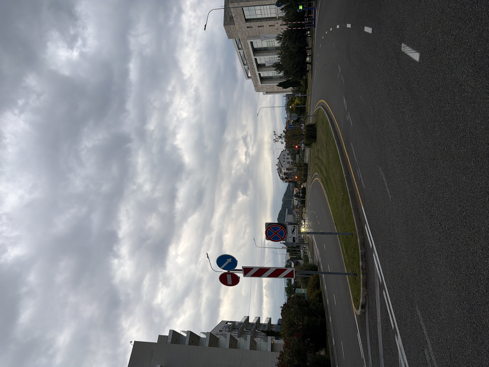 The image shows a street scene in Sirius, Russia, on a cloudy day. The road is paved and has white lane markings. On the left side of the road is a modern, multi-story building with balconies. Further down the road, on the right, is a large, contemporary building with a facade of light-colored stone and glass. Several streetlights are visible, as well as traffic signs. One sign depicts a red circle with a diagonal line through it (prohibiting entry) and a blue arrow indicating a left turn. Another sign shows a red cross within a blue circle, indicating no parking, and an image of a tow truck. Some trees and bushes are present along the roadside. There are a few cars visible on the road, and the sky is overcast with heavy clouds.