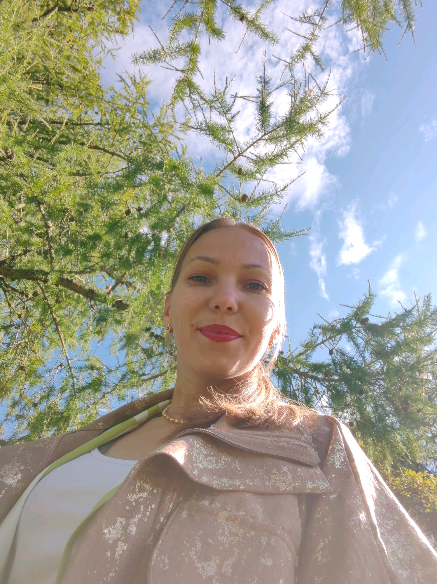 The image is a low-angle, outdoor selfie taken on a sunny day in the city of Chukhlem, Russia. A woman is centered in the frame, looking up at the camera. She has fair skin, light brown hair, and is wearing red lipstick and a gold necklace. She is also wearing a tan jacket with a metallic sheen over a white top, and a green collar is visible. She is positioned in front of a large tree with green needles, with a blue sky and scattered white clouds visible through the branches. The lower portion of the image shows more of her jacket and the tree's branches.