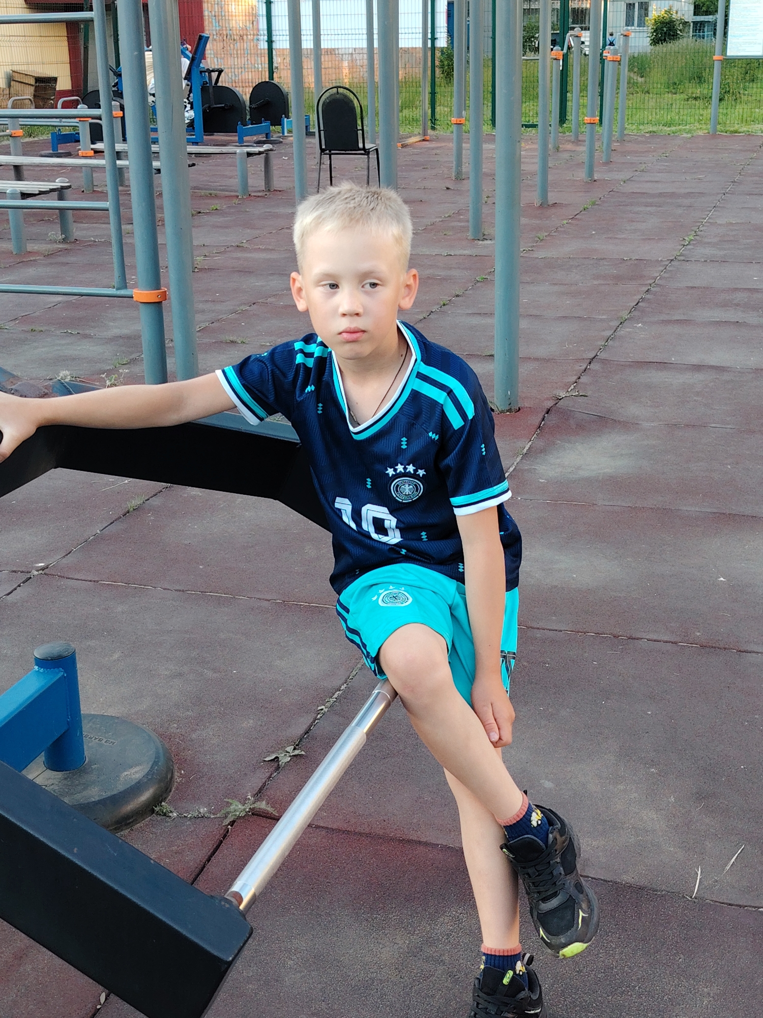 The image shows a young boy with blond hair sitting on a metal bar in an outdoor gym area. He is wearing a dark blue soccer jersey with light blue stripes on the sleeves and shorts. The jersey has the number "10" on the front and a crest with four stars. He is also wearing black athletic shoes and dark blue socks with a yellow pattern. The boy is looking towards the left of the frame with a neutral expression.
The gym area appears to be located in a park or public space. There are various pieces of exercise equipment visible in the background, including parallel bars, benches, and poles. The ground is covered with a reddish-brown rubberized surface, typical of playgrounds and outdoor gyms. In the far background, there are buildings and a green fence.
Based on the surrounding environment and the presence of outdoor gym equipment, this scene is consistent with a public park or sports facility in the city of Chaikovsky, Russia. The time of day appears to be late afternoon or early evening, given the lighting.