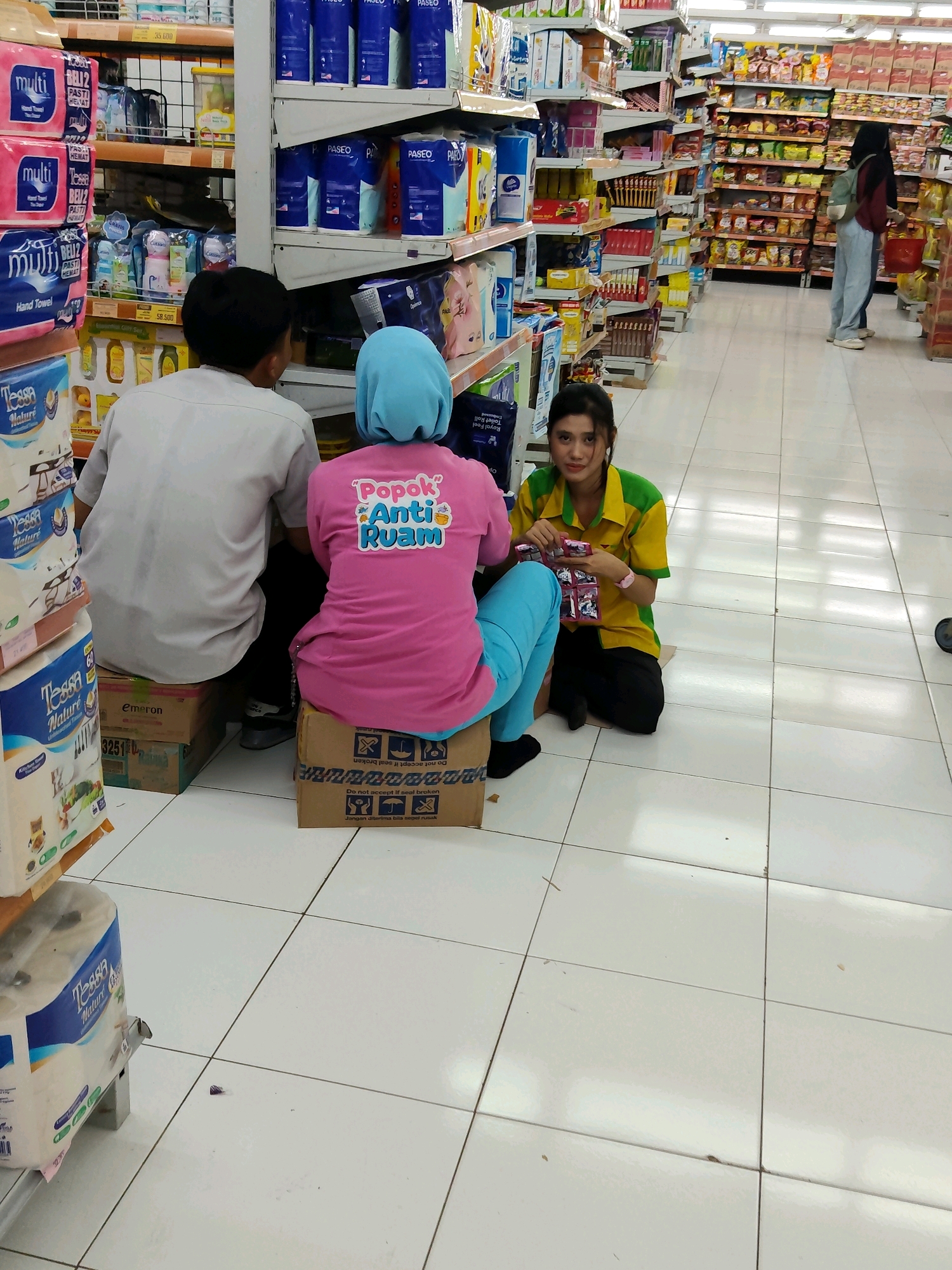 The image depicts a scene inside a well-stocked supermarket in Purwakarta, Indonesia. Three individuals are the primary subjects. In the foreground, a man and a woman are seated on cardboard boxes. The man, viewed from behind, wears a light gray collared shirt and dark pants. The woman next to him wears a pink t-shirt with a graphic design on the back that says "Popok Anti Ruam" (Diaper Rash Cream) and blue leggings. A second woman, likely an employee, is seated on the floor to the right of the couple, wearing a yellow and green collared shirt and black pants. She is holding several small packages, possibly for sale. The environment is characteristic of a supermarket aisle, with shelves stocked with various products including paper goods, packaged foods, and personal care items. The floor is tiled with white squares. A woman in a hijab and backpack can be seen in the background, browsing a different aisle. The lighting appears to be standard fluorescent supermarket lighting.