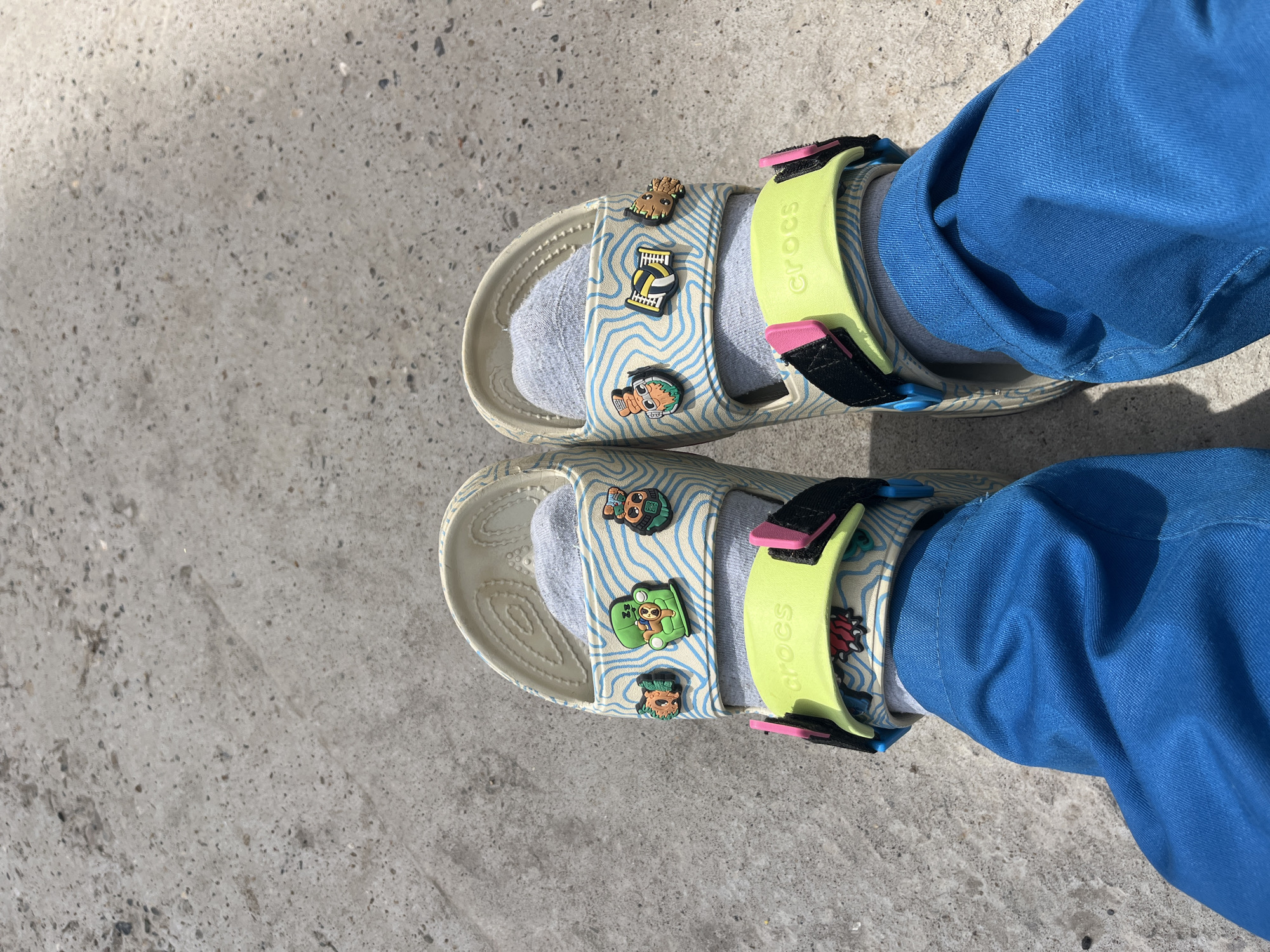 A downward shot shows a person wearing blue pants, grey socks, and patterned Crocs sandals. The sandals are adorned with cartoon character Jibbitz. The background consists of grey concrete. The location and the name of the city Chaikovsky, Russia cannot be determined based on the image.