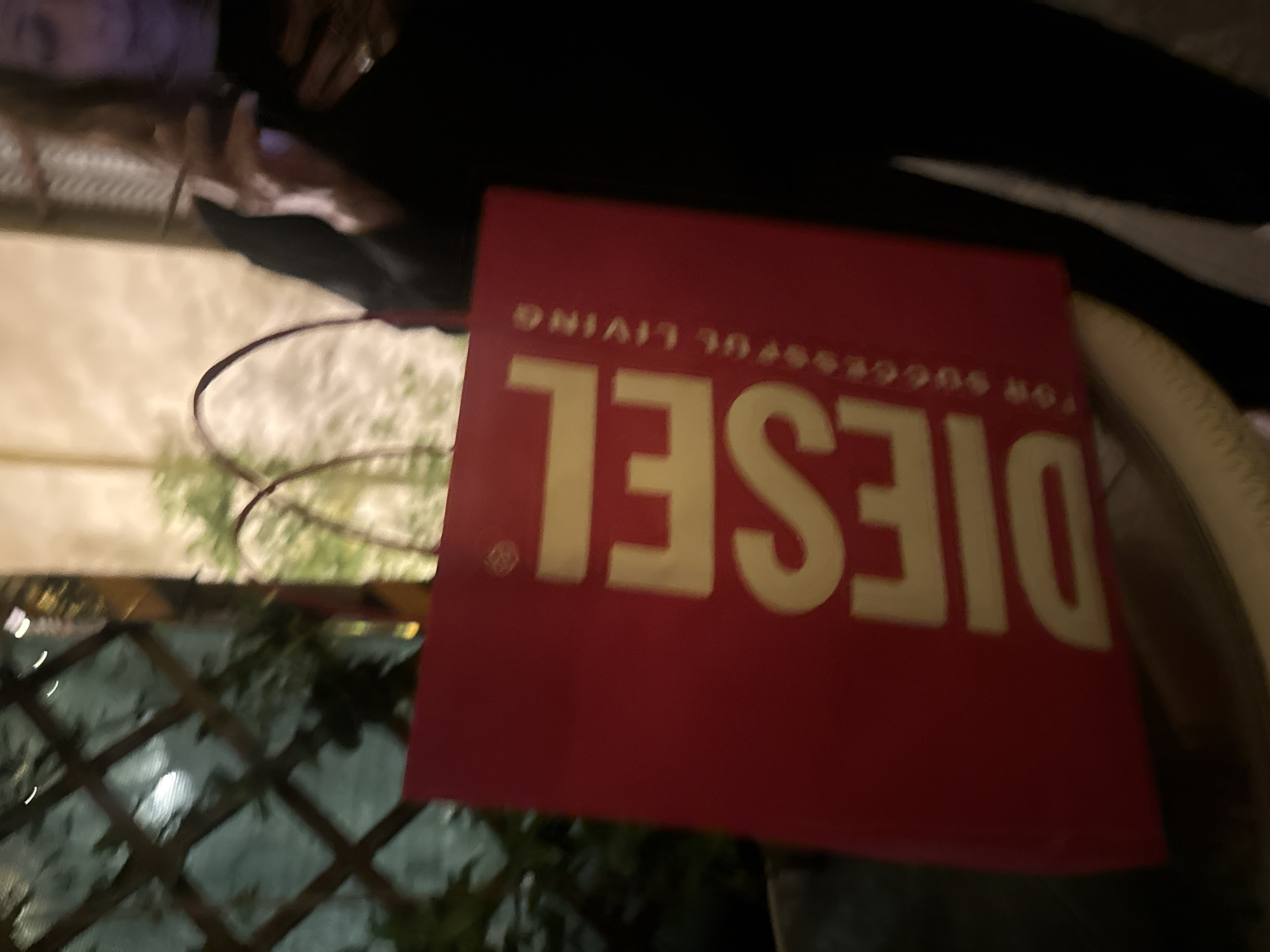 A red Diesel brand shopping bag is prominently displayed against a blurred background that includes a lattice-like structure covered with greenery. The bag is facing slightly to the right, with the "DIESEL FOR SUCCESSFUL LIVING" logo printed in large, beige, mirrored letters. Parts of a person and a white, textured object are partially visible in the top portion of the photo. The setting appears to be an outdoor space, likely a cafe or restaurant, as suggested by the environment and items in the background. The city of Milan, Italy, is not discernible from this image.
