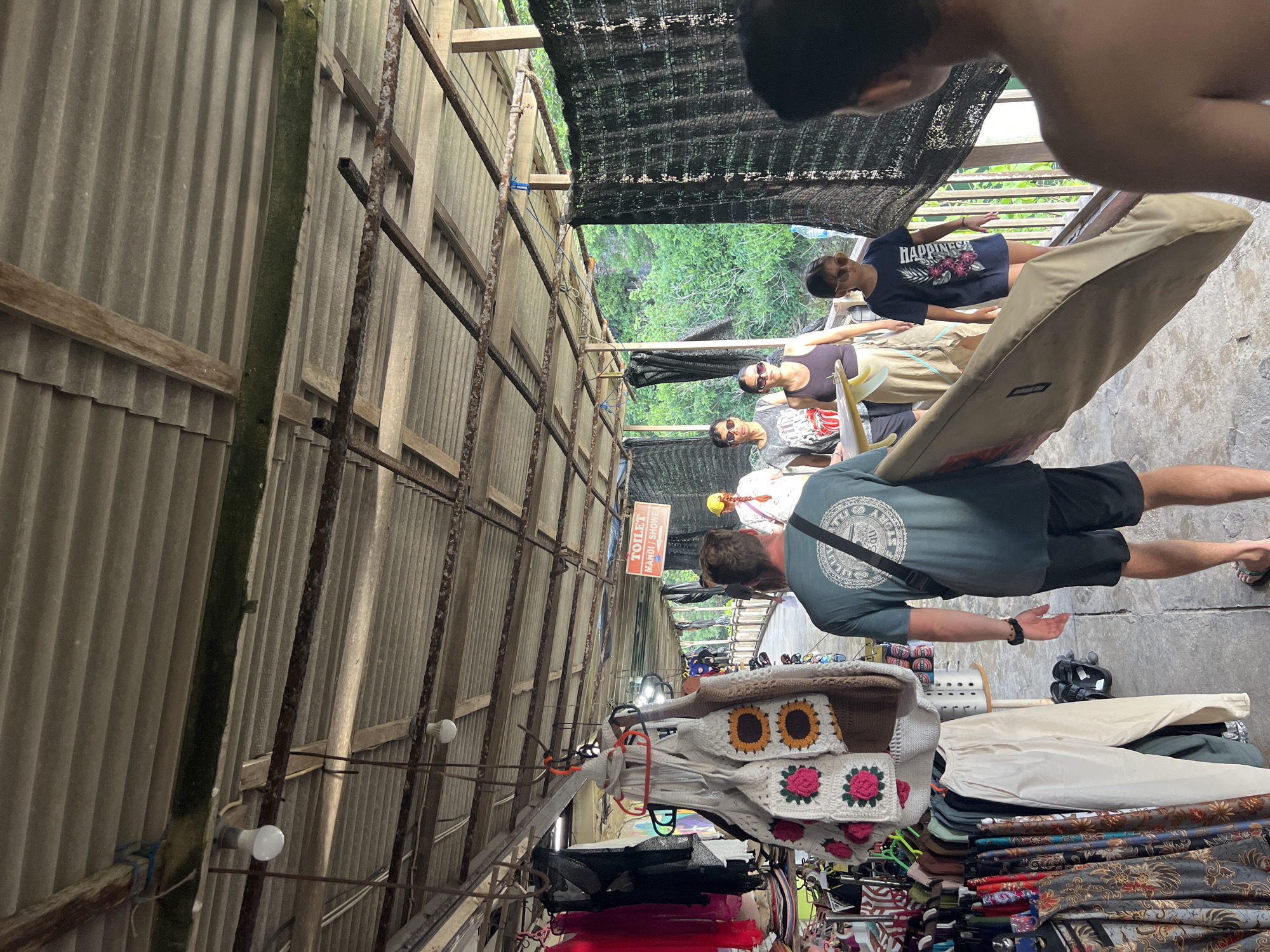 The image depicts an indoor scene in Kuta Selatan, Indonesia, within a market or shopping area. The structure appears to be built with corrugated metal siding and a metal framework. People are present, including individuals browsing the stalls. Various merchandise items are displayed, such as clothing and fabrics. Overhead lighting is visible. A sign with the words "TOILET" and "MANDI / SHOWER" is hanging in the market. The environment appears to be somewhat enclosed and cluttered with the displays.