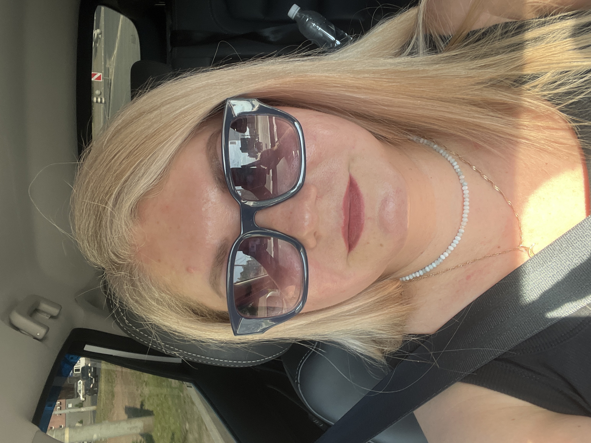 A light-skinned woman with blonde hair is wearing sunglasses and a black seatbelt inside a car. She is wearing a layered necklace consisting of white beads and a gold chain. A water bottle is visible in the background behind her head. Through the car window, buildings and greenery are seen. A street sign with a red symbol is also visible. This is probably near the city of Izhevsk, Russia.