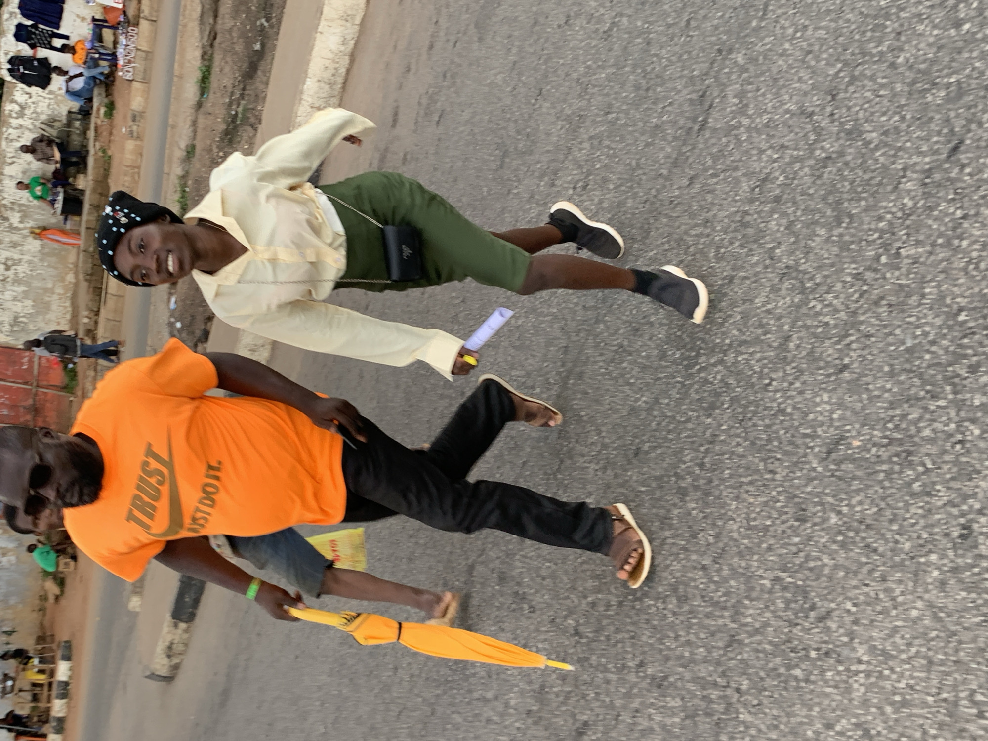 In Jos, Nigeria, two individuals walk on a paved road. A man wearing a bright orange t-shirt with the text "TRUST JUST DO IT" and sunglasses walks ahead, carrying a closed yellow umbrella. He is also wearing dark pants and brown sandals. To his right, a woman in a light yellow button-down shirt and a dark green skirt walks alongside him. She has a black headscarf with white polka dots and a small black purse with a chain strap slung across her body. She is wearing black sneakers with white soles. In the background, other people and market stalls are visible along the roadside.