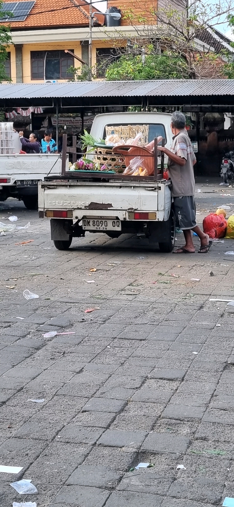 In Denpasar, Indonesia, a man in a light brown, short-sleeved shirt and grey shorts is standing next to the open tailgate of a white Suzuki pickup truck. The truck's bed contains several items, including woven baskets filled with what appear to be food items and green vegetables. In the background, a second white Suzuki pickup truck is visible with a license plate that reads "DK 8799 CV". The ground is paved with grey interlocking stones and littered with various pieces of trash, including plastic cups, wrappers, and leaves. A red and yellow plastic bag are also visible near the truck. The scene appears to be an outdoor market or roadside area.