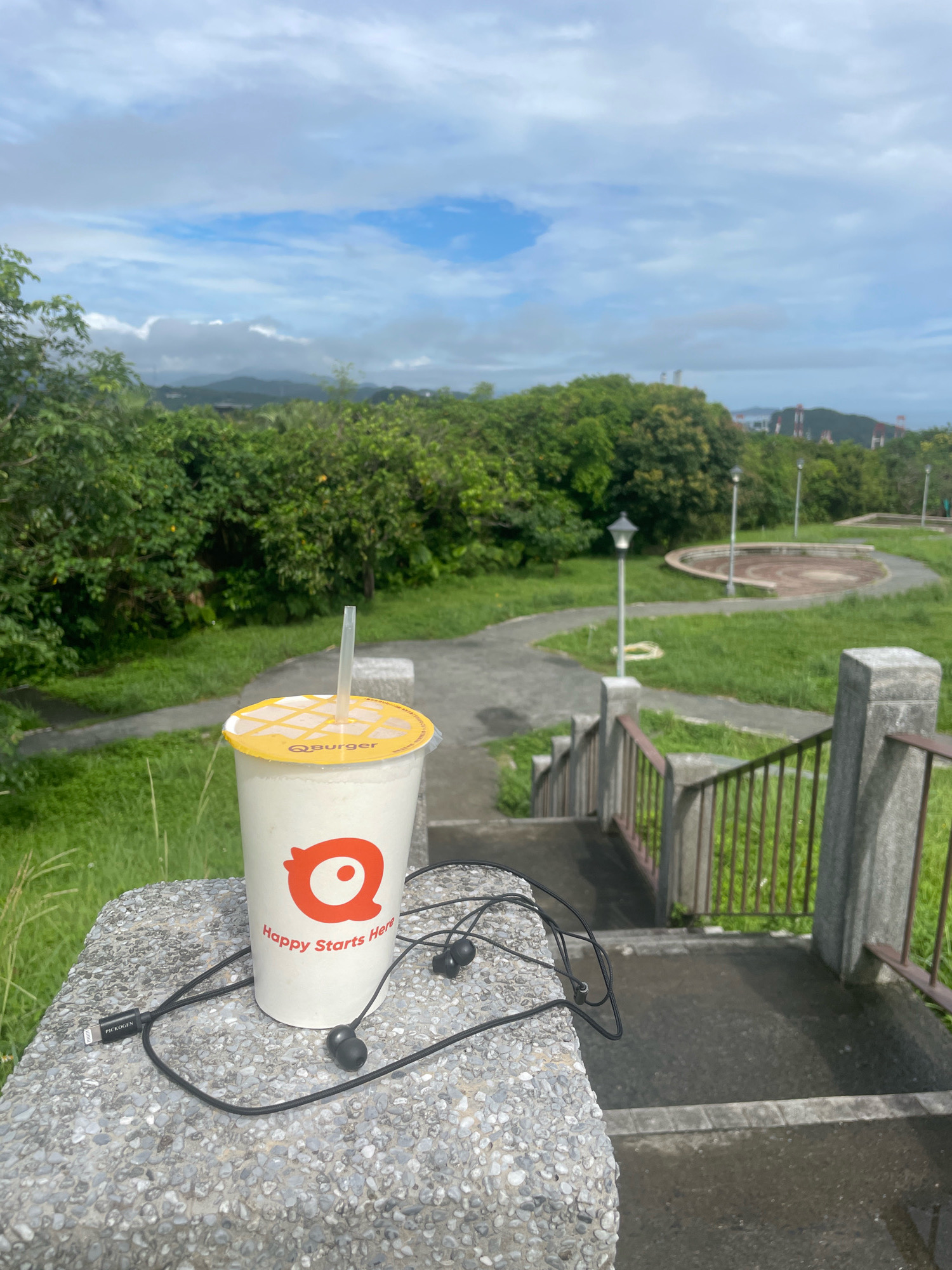 A white paper cup with a yellow lid and a straw sits on a speckled grey concrete ledge. The cup has an orange logo and the text "Happy Starts Here" printed on it. Black wired earbuds are coiled next to the cup. The concrete ledge overlooks a park with green grass, trees, a paved path, and a set of concrete stairs with metal railings. In the background, hills and a cloudy sky are visible. The location is likely near the Keelung Coastal Park in Keelung, Taiwan, given the distinctive landscape and vegetation.