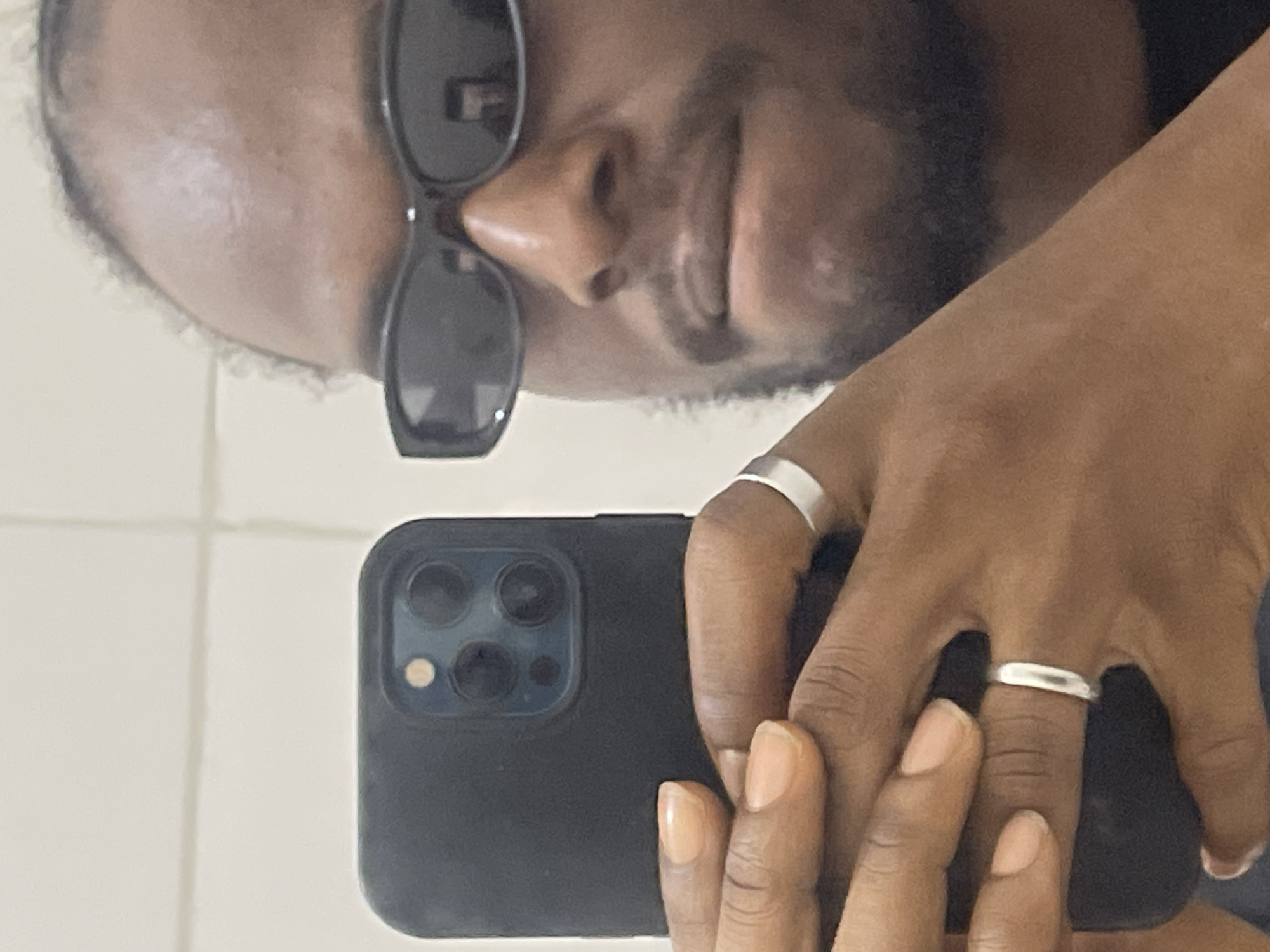 The image captures a mirror selfie of an individual, likely male, from the mid-chest up. The person has dark brown skin, a trimmed beard and mustache, and dark hair with some visible grey strands near the forehead. They are wearing black rectangular sunglasses with dark lenses, which obscure their eyes and reflect a blurry image of the immediate surroundings, including part of their own head. The subject is holding a black iPhone Pro model smartphone in a matte black case with both hands, positioned to capture the reflection. The phone's rear camera module, featuring three prominent dark camera lenses and a smaller flash/sensor module, is clearly visible. Two simple silver-toned rings are worn: one on the left ring finger and another on the right middle finger. The fingernails on both hands are short and coated with a light peach or nude-colored polish. The background consists of a light beige or off-white tiled wall with visible grout lines, suggesting an indoor setting. The image provides no discernible visual information to establish its location in Jos, Nigeria.