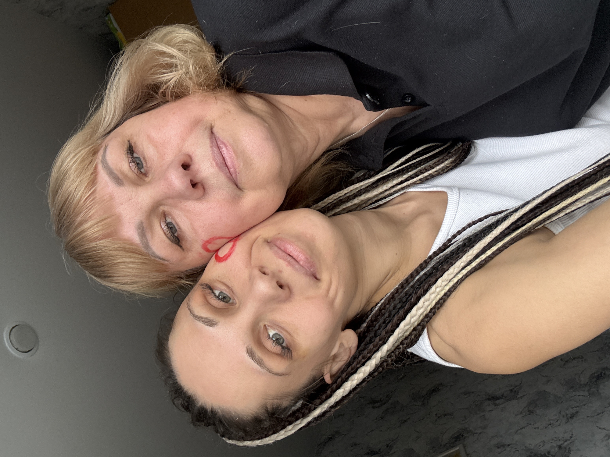 A close-up, top-down portrait features two women, likely a mother and daughter, captured in an indoor setting in Rusanovo, Russia. The image is oriented sideways, with both women looking up towards the camera, their heads close together in an affectionate pose.
The older woman, positioned on the left, has blonde hair with bangs and is wearing a black collared shirt. She has a gentle smile and light eye makeup. Her head is nestled against the younger woman's cheek.
The younger woman, on the right, has dark hair styled in long braids with prominent light-colored extensions. She is wearing a white top, possibly a tank top. A distinctive red heart shape is drawn on her left cheek, near her eye, adding a playful or symbolic element to the image. Both women share soft, pleasant expressions, conveying a sense of warmth and closeness.
The background is a plain, light gray surface, suggesting a wall or ceiling, with a dark circular fixture visible behind the younger woman. The lighting is bright and even, indicating an indoor daytime shot. The scene suggests a personal moment, possibly a selfie taken to celebrate their bond. There is no visible text in the image.
