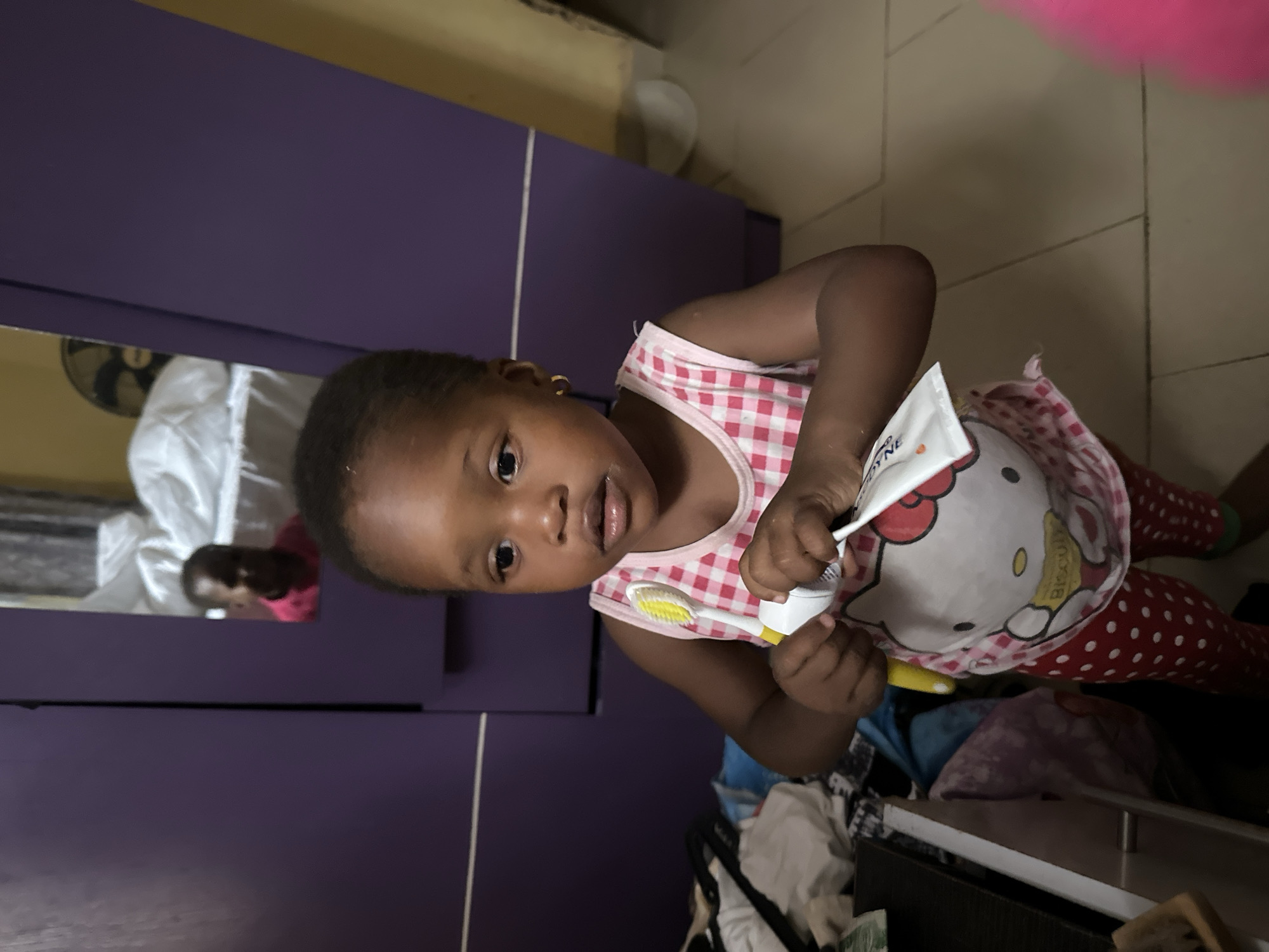 A young Black child, likely a toddler, is standing in what appears to be a bedroom in Narayi, Nigeria. The child is holding a tube of toothpaste and a toothbrush, with the bristles of the toothbrush touching the opening of the toothpaste tube, suggesting an attempt to apply toothpaste. The child is wearing a pink and white checkered tank top with a Hello Kitty graphic on the front and red polka dot leggings. Visible in the background is a large purple wardrobe with a mirror. The mirror reflects another child, also wearing a pink shirt. The room appears cluttered, with clothing and other items visible near the wardrobe. The floor is tiled. A fan is visible in the background, mounted on the wall.