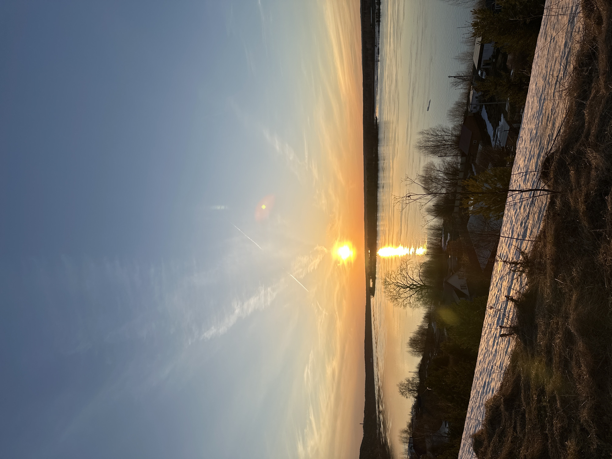 The image depicts a sunset over a body of water, likely a river or lake, in the vicinity of Ольховка, Russia. The sun is positioned on the horizon line, its light reflecting off the water's surface. Contrails are visible in the sky. Buildings and trees are on the bank of the water, indicating a populated area. A patch of snow is visible in the foreground.