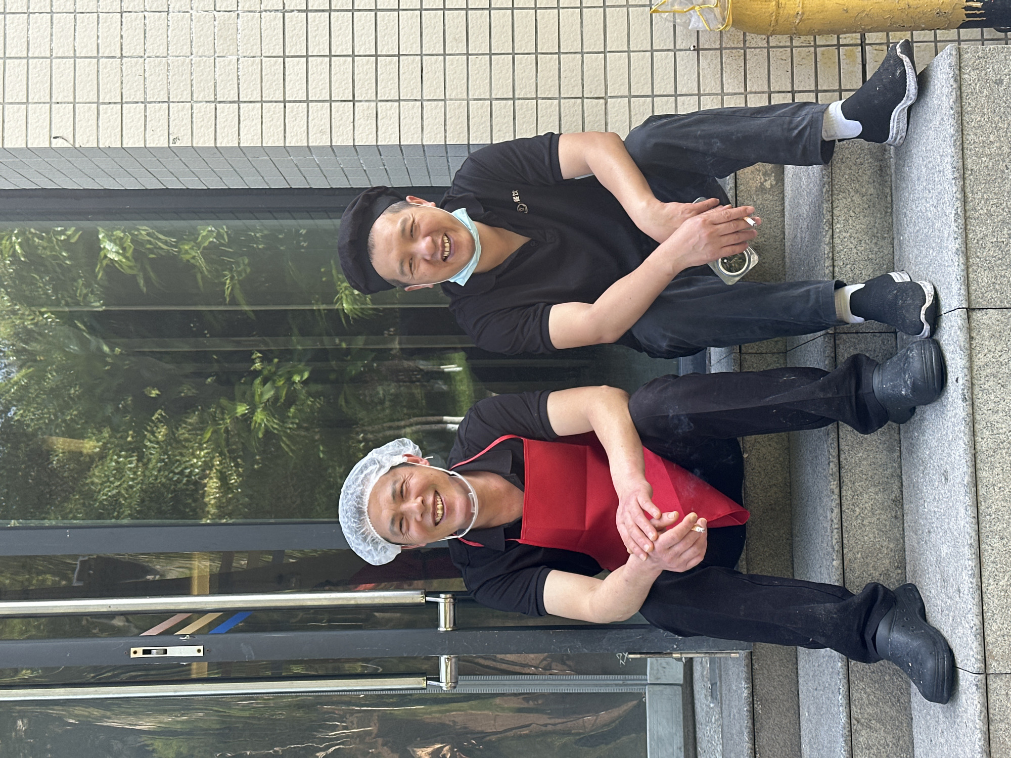 Two men, likely kitchen staff, are captured sitting on concrete steps outside a building, possibly an eatery, in Jiangjin, China. Both are smiling and looking towards the camera, appearing relaxed and taking a break.
The man on the upper step wears a black beret, a black polo shirt, black pants, and black shoes with white socks. A light blue face mask is pulled down under his chin. He holds a lit cigarette in his right hand and a small clear container, possibly used as an ashtray, in his left. On his polo shirt, a logo with Chinese characters "悦文" (Yuè Wén) is visible.
The man on the lower step wears a white bouffant cap (hairnet), a black polo shirt, a red apron, black pants, and black clogs. He also holds a lit cigarette in his right hand.
Behind the men, there is a light-colored tiled wall. To their left, large glass windows reflect lush green foliage, suggesting trees or bushes nearby. Metal frames and a latch mechanism are visible on the window structure. In the background, on the right side of the image, a yellow pipe or bollard is partially visible. The scene is well-lit, indicating daytime. The men's expressions convey contentment and friendliness.