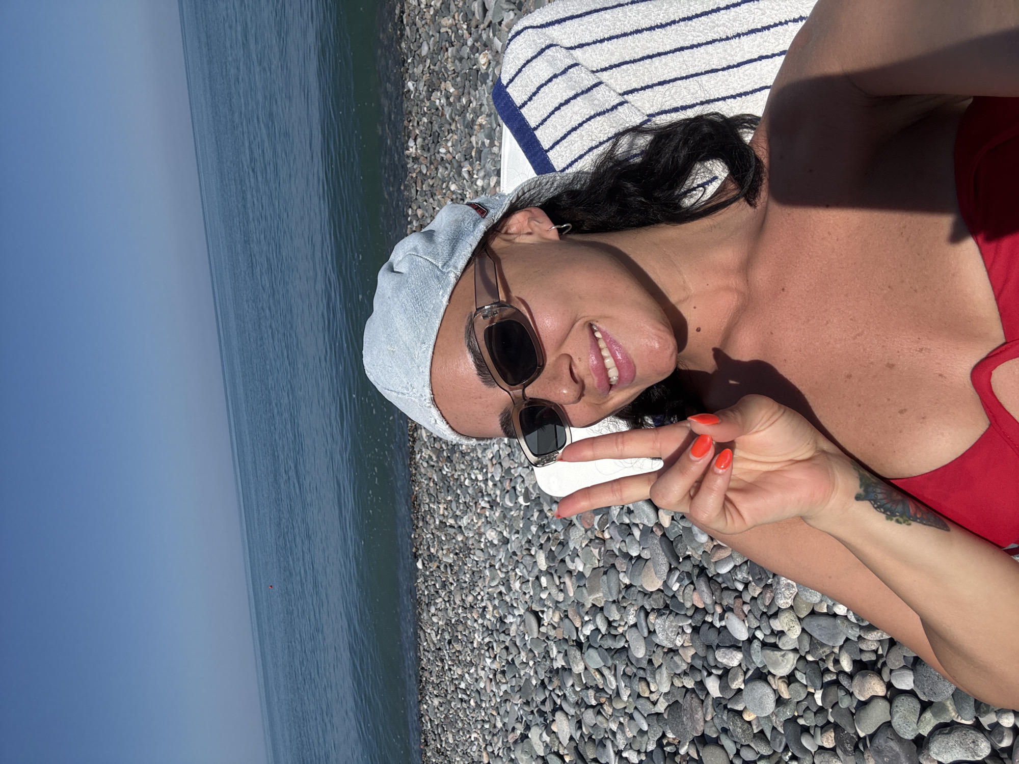 A woman is lying on a beach chair on a pebble beach in Batumi, Georgia, posing for a selfie. She is wearing a red bikini top and a light blue cap, with sunglasses on. Her hand is raised, displaying a peace sign with bright orange nail polish. The sea is visible in the background, with a clear blue sky above. The beach is covered in smooth, grey pebbles.