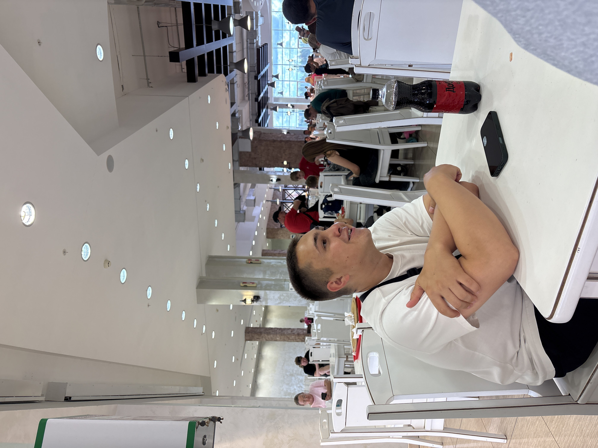 The image depicts a young man sitting at a white table in what appears to be a food court or cafeteria. He is wearing a white t-shirt and black pants, with his arms crossed. A dark-colored smartphone and a bottle of dark soda are on the table in front of him. In the background, other patrons are seated at tables, and the environment features a high ceiling with numerous lights, columns, and brick-patterned walls. The overall impression is that of a casual, public dining area. It's not possible to determine the specific location within the city of Perm, Russia from the image alone.