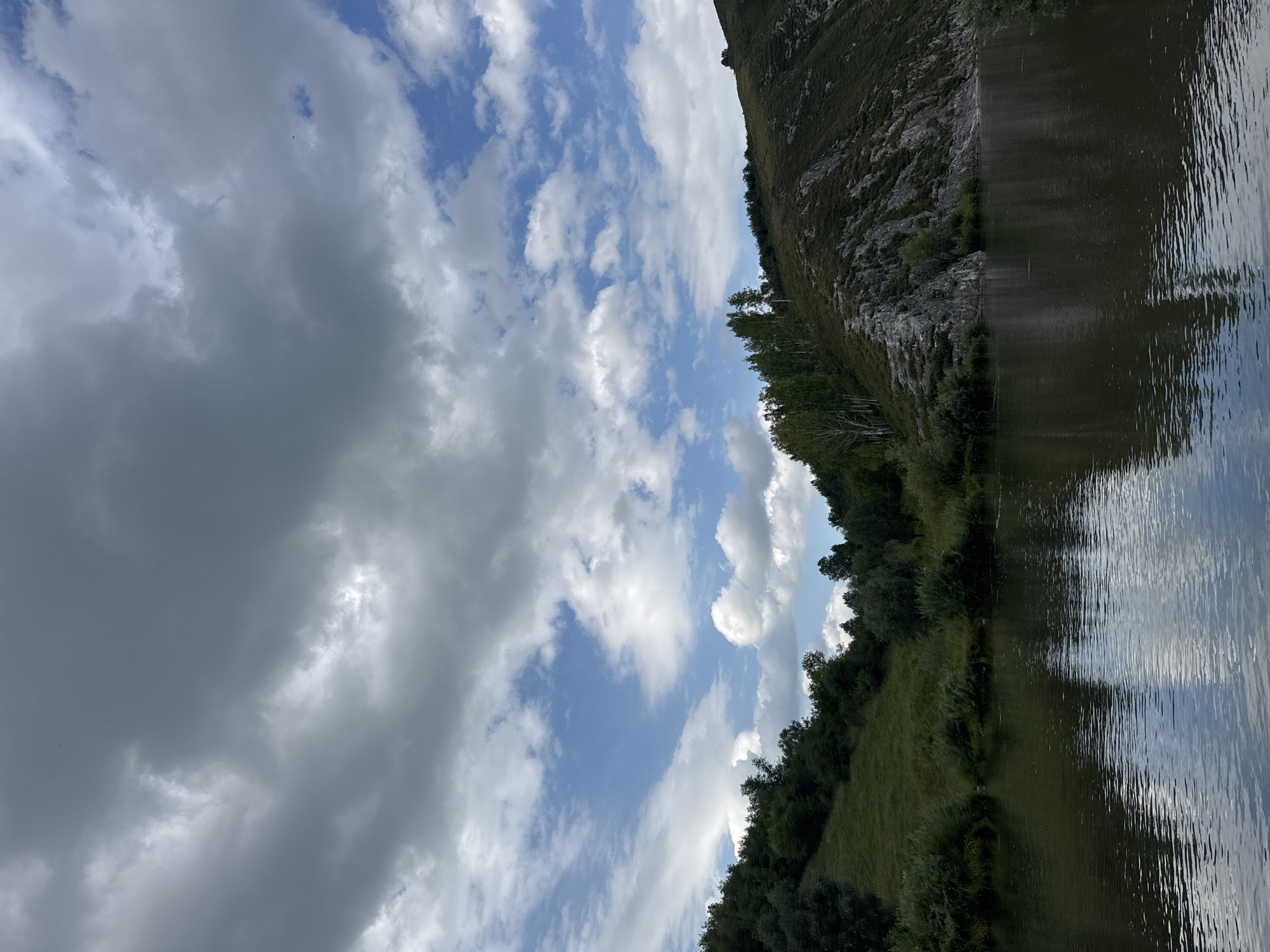 The image depicts a natural landscape with a river in the foreground and rolling hills covered in greenery on the opposite bank. The sky is filled with dramatic clouds, with patches of blue sky visible between them. The water in the river is dark and reflects the surrounding environment. The hills are densely wooded in some areas and rocky in others, with a path or trail visible on one of the rocky slopes. The scene suggests a tranquil, outdoor setting, possibly a park or natural reserve within or near the city of Soloneshnoye, Russia.