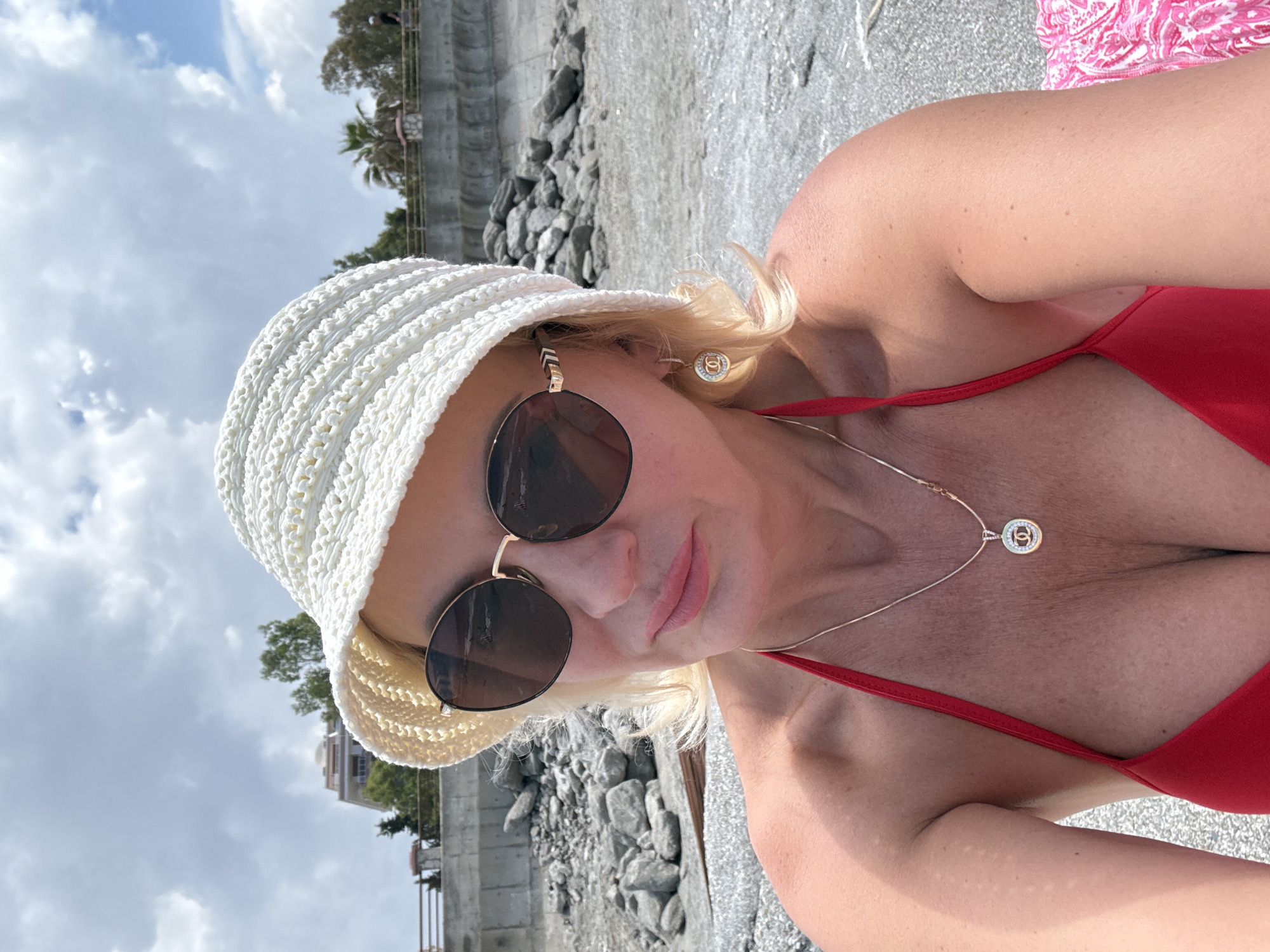 A woman with blonde hair, wearing sunglasses and a white hat, is posing for a selfie. She is wearing a red top and a necklace with a circular pendant. The background includes a rocky area, a wall or barrier, trees, buildings, and a sky with clouds. The location appears to be a beach or coastal area.