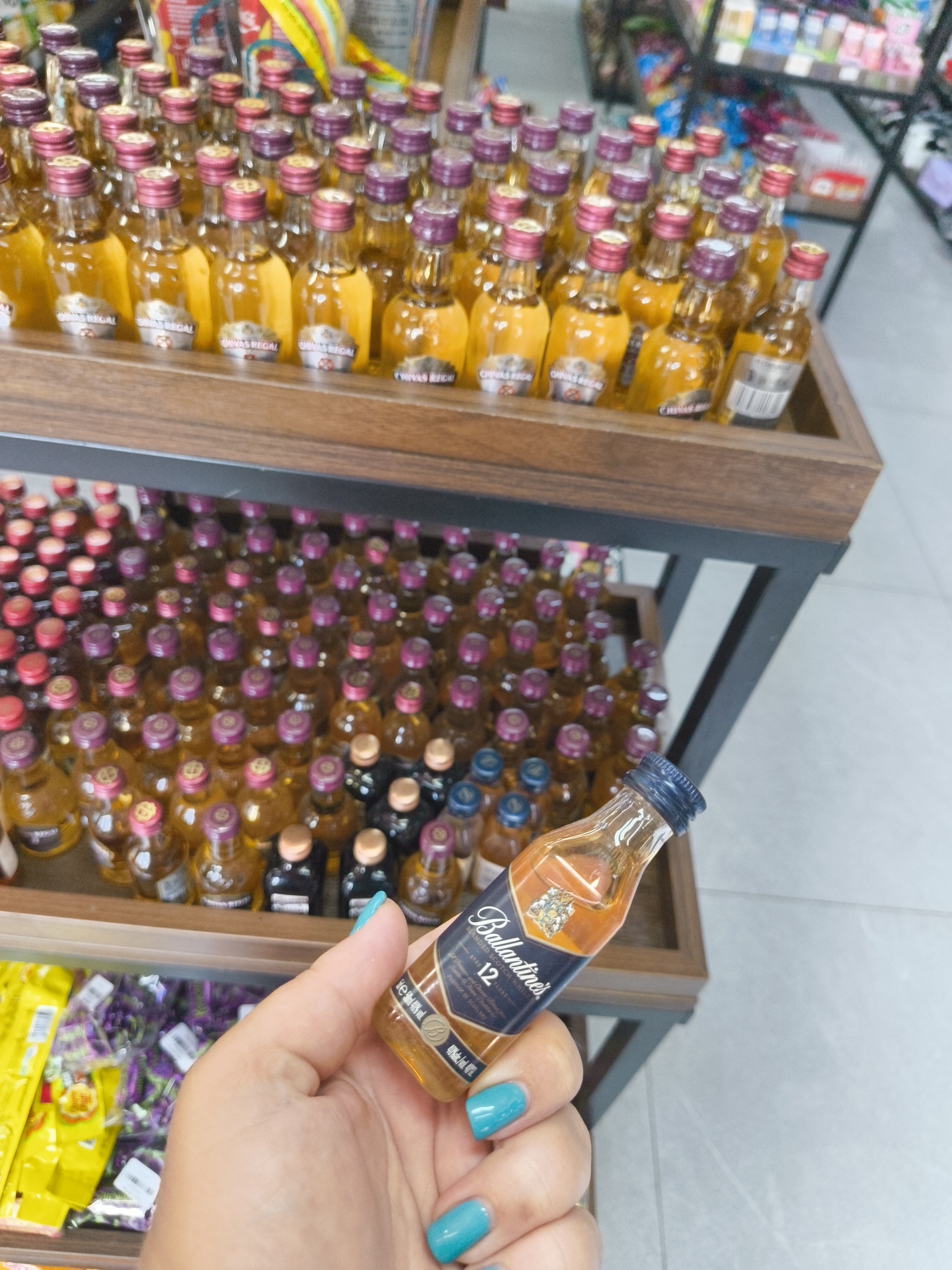 The image shows a hand holding a miniature bottle of Ballantine's 12 Year Old Scotch Whisky. The bottle has a blue cap and a dark blue label with gold lettering. The background shows a shelf filled with many other miniature bottles of alcohol, mostly whisky, arranged in rows. There are also some yellow and purple packaged items visible on a lower shelf. This scene appears to be in a retail store, possibly a duty-free shop or a convenience store, given the variety of small bottles of spirits. The overall impression is that of a display of miniature alcoholic beverages for sale. The lighting is bright and even, suggesting an indoor setting. The image was taken from a slightly elevated angle, looking down at the hand and the bottle.