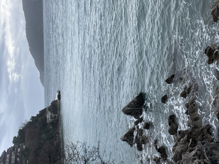 The image displays a coastal scene in Budva, Montenegro, under an overcast sky. The foreground consists of a rugged, dark gray rocky shoreline with white sea foam indicating minor wave activity. Bare tree branches are visible on the left.
The midground features the Adriatic Sea, characterized by dark teal-blue water with a rippled surface. Two prominent, triangular dark rocks protrude from the water near the shore, with a thin, light-colored line, consistent with a rope or wire, descending from the apex of the rightmost triangular rock into the water.
In the background, a steep, tree-covered hillside on the left supports a large, multi-tiered building with multiple balconies, likely a hotel or residential complex. Below this structure, at the waterline, a smaller, white, flat-roofed building or platform is situated on a rock formation. A distant, dark green mountainous landmass or island is visible across the horizon on the right. No individuals or specific human actions are depicted.
