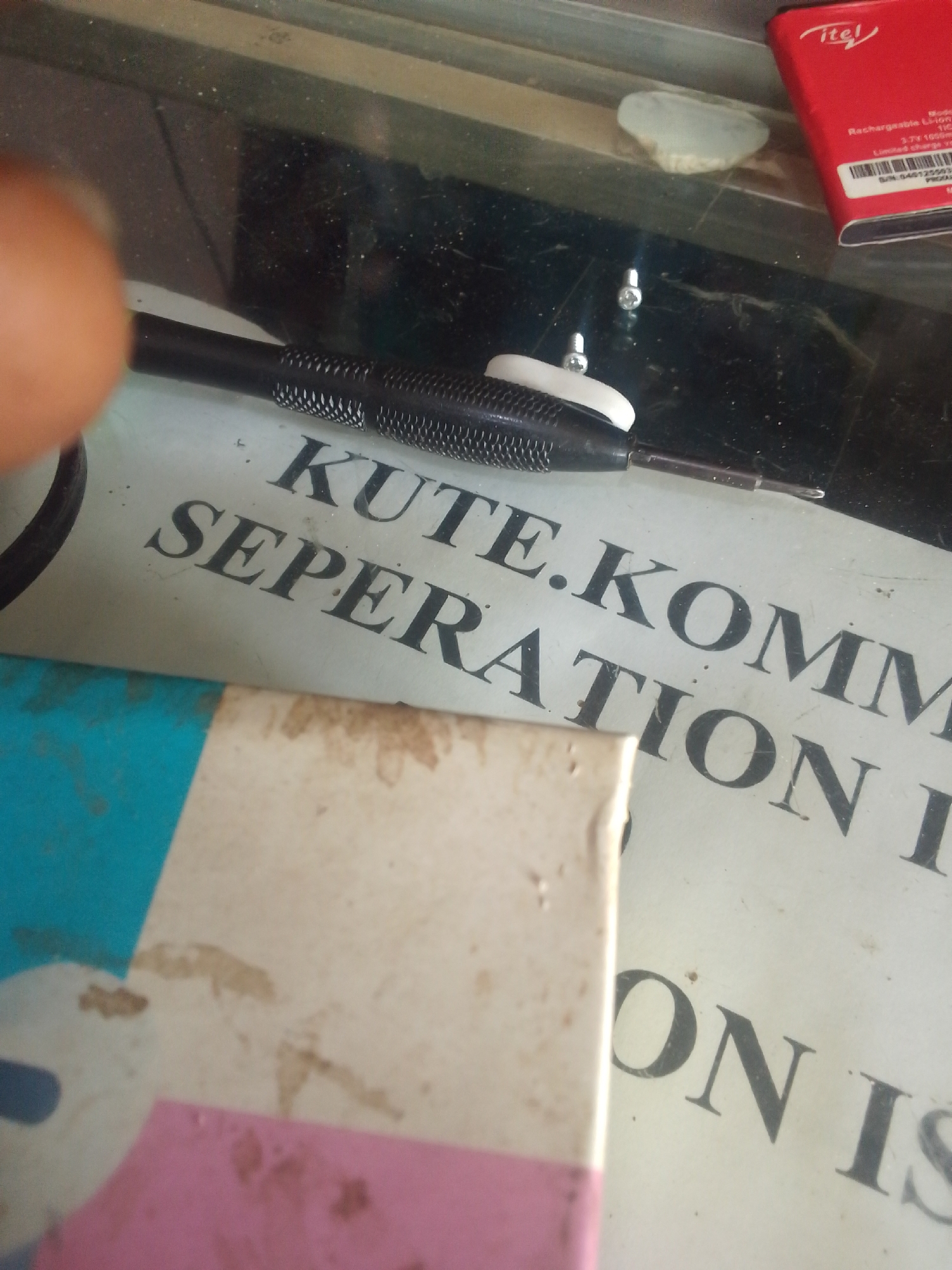 This image depicts a dusty work surface, identified as being in the city of Jos, Nigeria. The primary visible objects include a black-handled precision screwdriver with a textured grip and a white plastic collar near its tip, positioned centrally. To the right of the screwdriver, two small silver screws are visible on a dark, reflective surface that appears to be glass. Further to the right, a red rectangular Itel-branded rechargeable Li-ion battery (3.7V, 1000mAh) rests on the same surface. Below the screwdriver, a partially obscured white sheet of paper displays black text, clearly reading "KUTE.KOMM SEPARATION I." In the lower left, a portion of a blue object is visible, and in the lower right, a rectangular pink and white object, possibly an eraser or small box, is present. A blurry, out-of-focus finger with light brown skin tone occupies the upper-left corner of the frame. The overall environment suggests a workspace, possibly an electronics repair shop, characterized by scattered dust and minor debris. No other individuals or specific actions are discernible beyond the objects themselves and the implied setting.