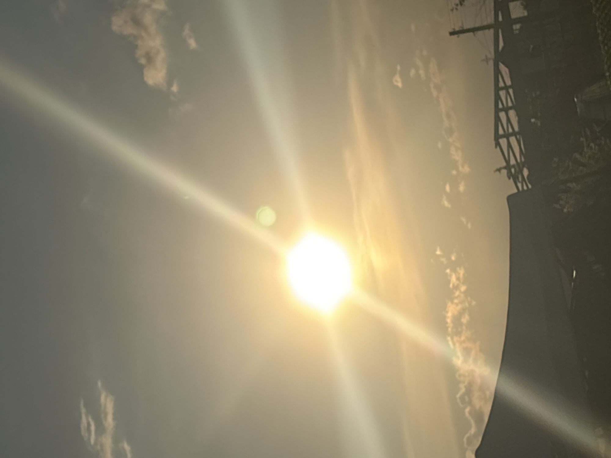The image depicts a bright, overexposed sky with the sun prominently visible, producing strong lens flares. There are faint clouds scattered throughout the sky. In the lower right corner, a dark silhouette of a building or structure is present. In the upper right corner, there's a power or telecommunication pole, which indicates the location of the urban environment. There is no information in the image that shows that this scene is in Kukorevo, Bulgaria.