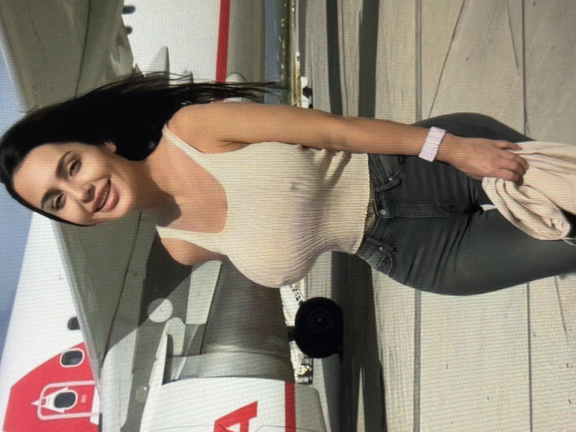 A woman with dark hair and light skin stands on an airport tarmac in front of a commercial airplane. She is wearing a light-colored tank top and dark jeans. The airplane has a red and white livery. The woman is holding a light-colored garment in her right hand. The image is taken from the chest down. There is no indication of the location being Shumen, Bulgaria.