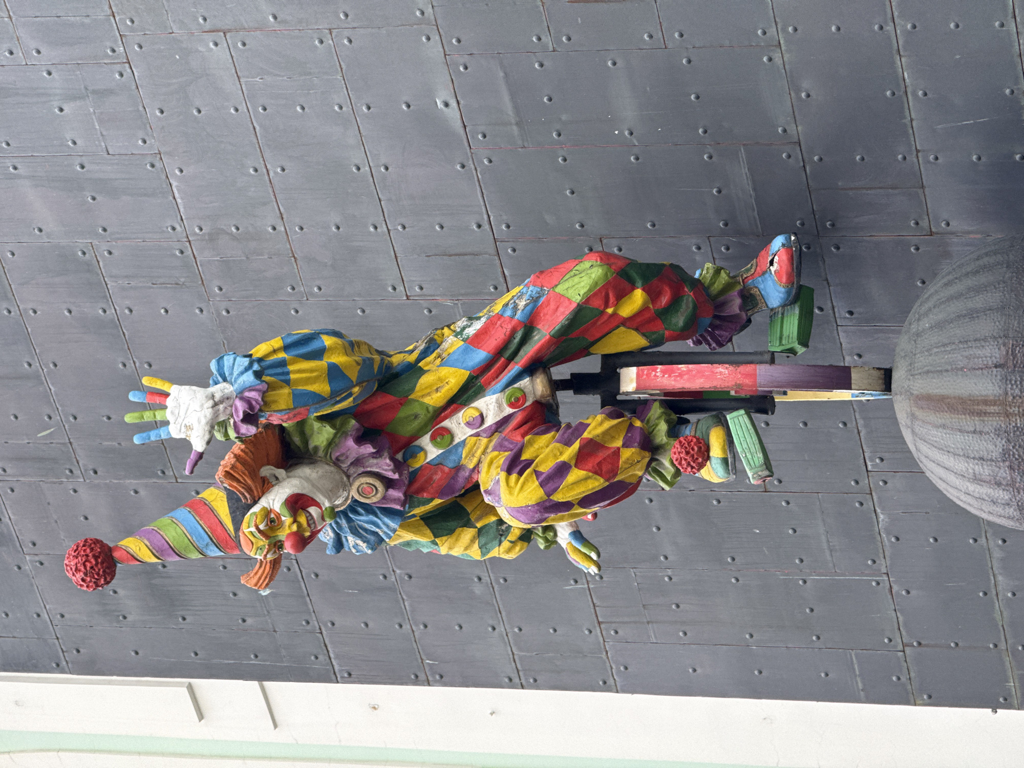 The image shows a sculpture of a colorful clown riding a unicycle, mounted on the exterior of a building. The clown is wearing a harlequin-patterned costume in vibrant red, yellow, blue, and green. It has a red ball on top of its striped conical hat and its face is painted with exaggerated features. The clown's hands are raised, with fingers spread. The unicycle is made of wood and painted, with a red wheel. The background is a dark, textured metal wall with visible rivets. To the left of the clown sculpture, a portion of a white building with a light green trim is visible. The base of the sculpture appears to rest on a dark, rounded dome-like structure. The sculpture is located in Moscow, Russia.