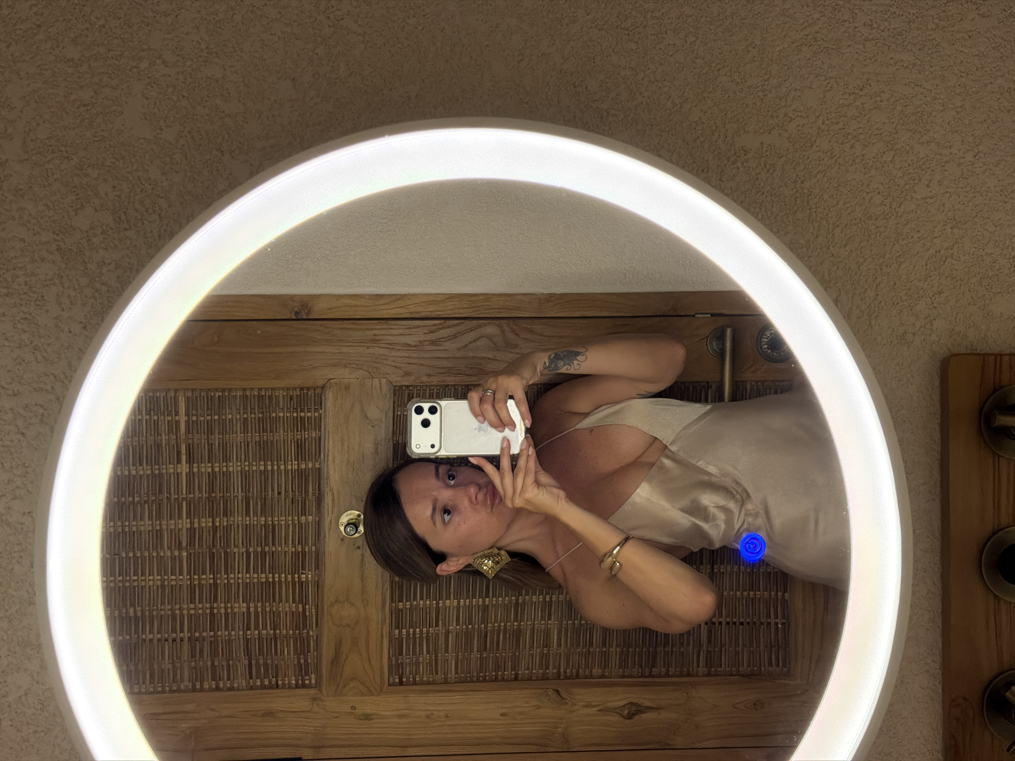 A woman is taking a mirror selfie in a dimly lit room. She is wearing a champagne-colored slip dress and large, gold geometric earrings. The mirror is round with a built-in LED light, and it reflects a wooden door with a woven panel and a brass doorknob. The walls appear to be textured and a light tan color. There are some objects visible on a shelf below the mirror, but they are out of focus. The woman's face is turned towards the camera, and she has her phone held up to take the picture. Her left arm is slightly raised, and her right hand is holding the phone. A tattoo is visible on her left forearm. The scene suggests a hotel room or a bathroom. The location is the city of Mengwi, Indonesia.