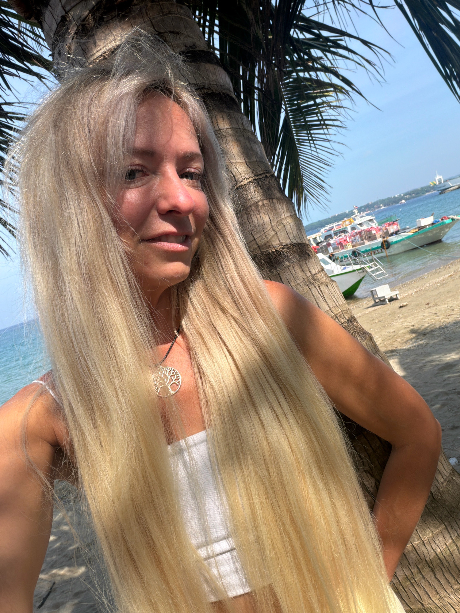 An outdoor, close-up, eye-level, medium shot captures a woman with long, blonde hair leaning against a palm tree on a beach. She is wearing a white top and a tree of life pendant. Behind her, several boats are anchored in the turquoise water near the shore, with a town visible in the distance. The scene is set during the day with clear skies and sunlight casting shadows. The location is likely a beach in Malay, Philippines, given the characteristic palm trees and boats.
