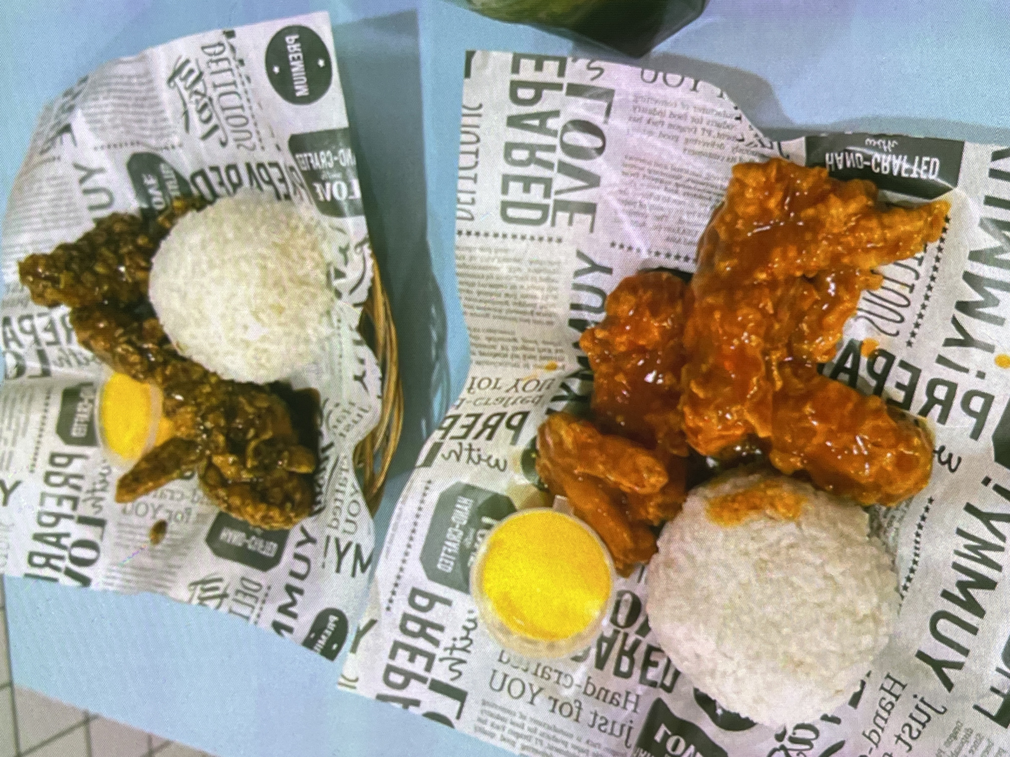 Two plates of fried chicken and rice are shown. Each plate is lined with patterned paper that appears to be newsprint. The chicken is coated in a dark glaze. A small cup of yellow sauce is on each plate. Both plates are on a light blue surface.